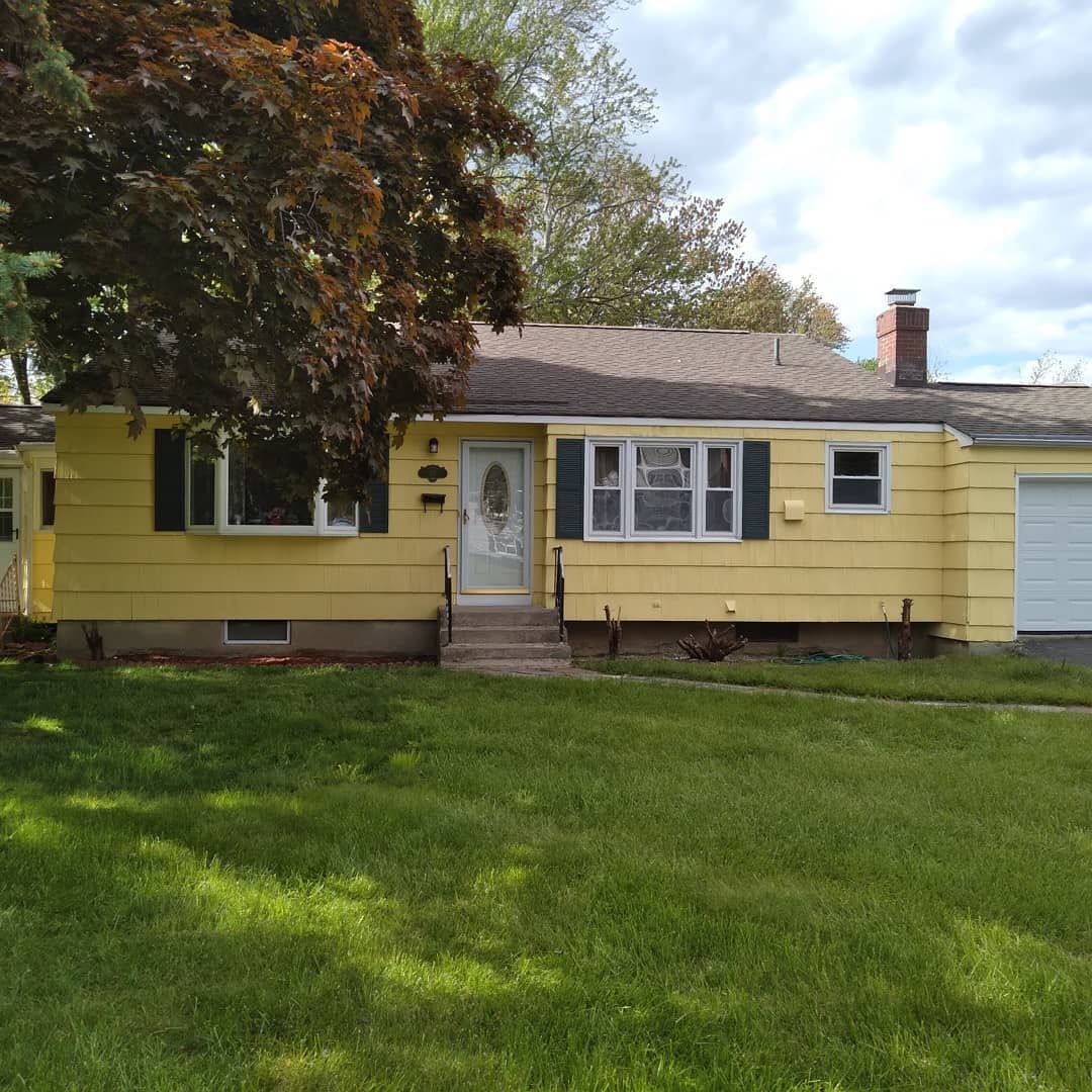 Yellow single-story house with white trim, front porch, and a large green lawn under a partly cloudy sky