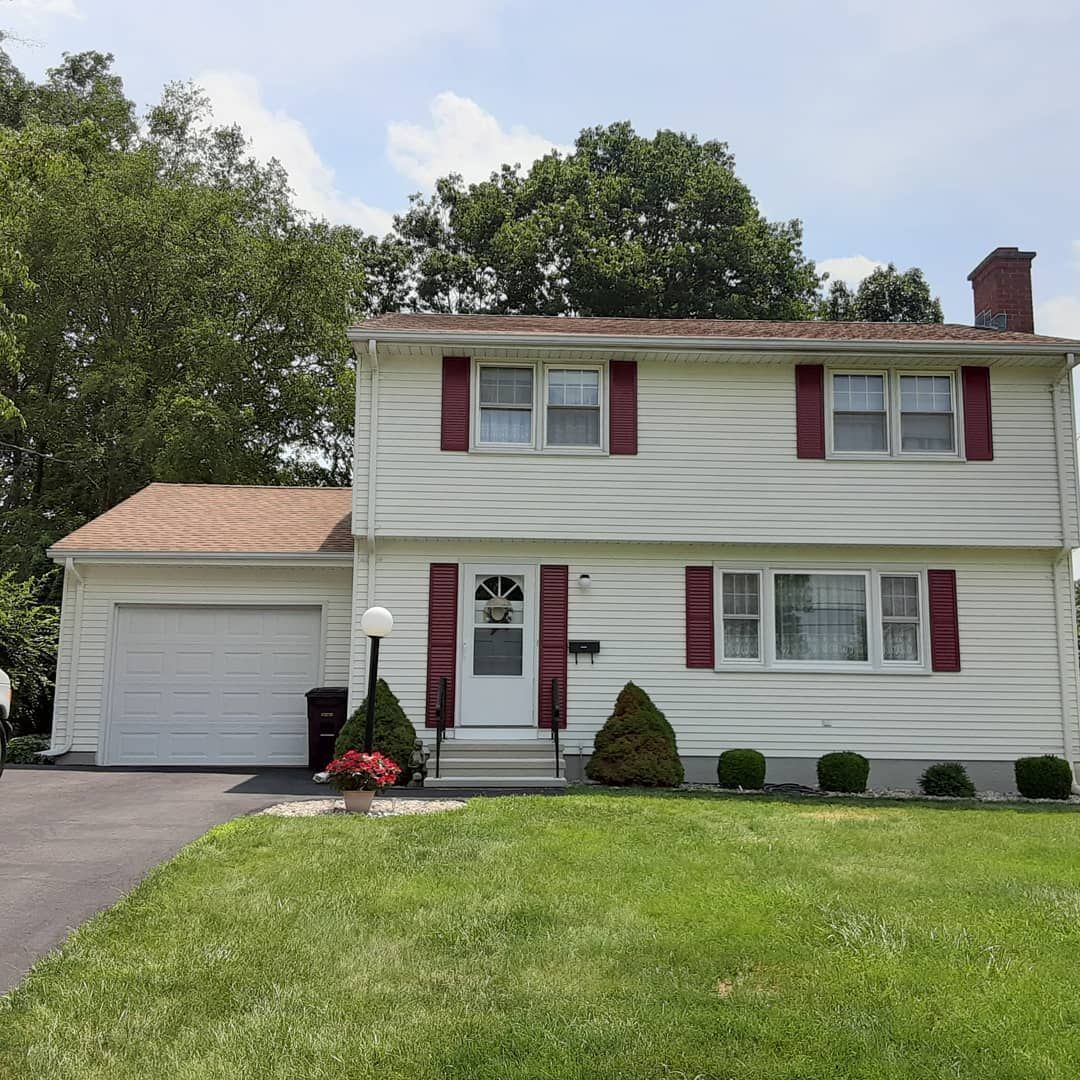 Suburban two-story white house with red shutters, attached garage, and a green lawn under a cloudy sky
