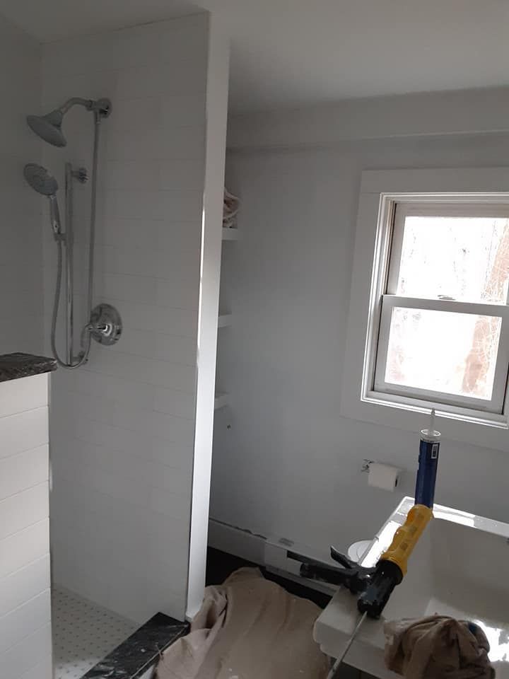 Small bathroom with shower and window; a person in beige pants is crouched near a sink.