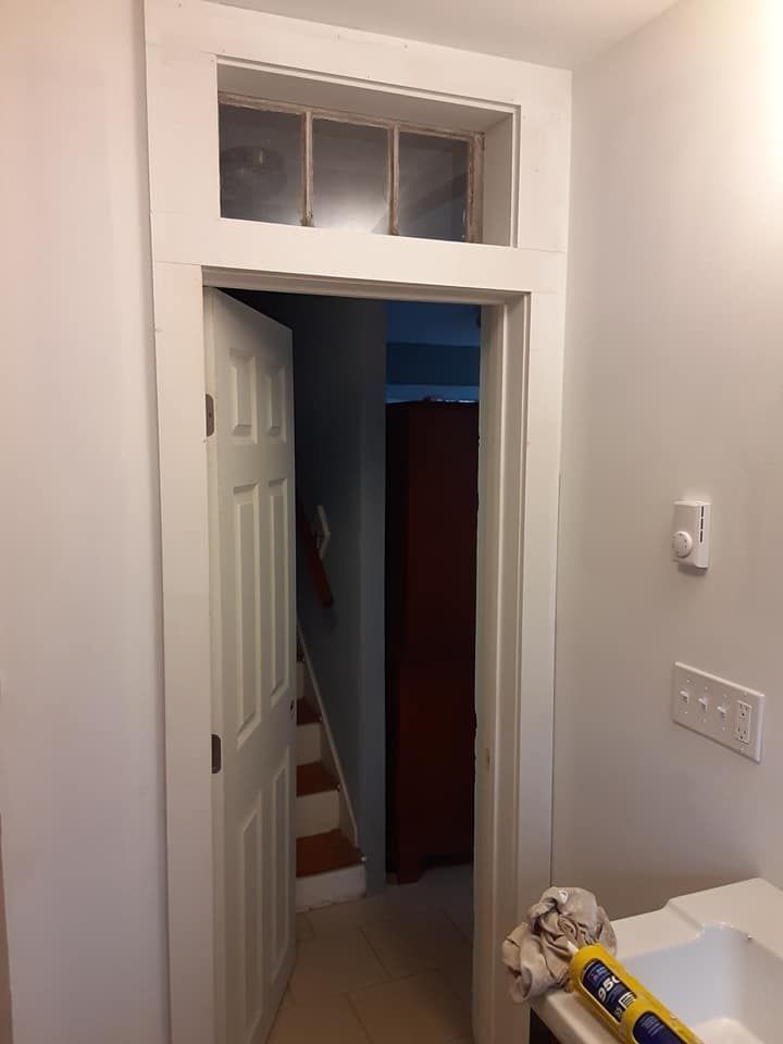 Narrow hallway with open white door, dark stairwell beyond, and a wall-mounted light switch.