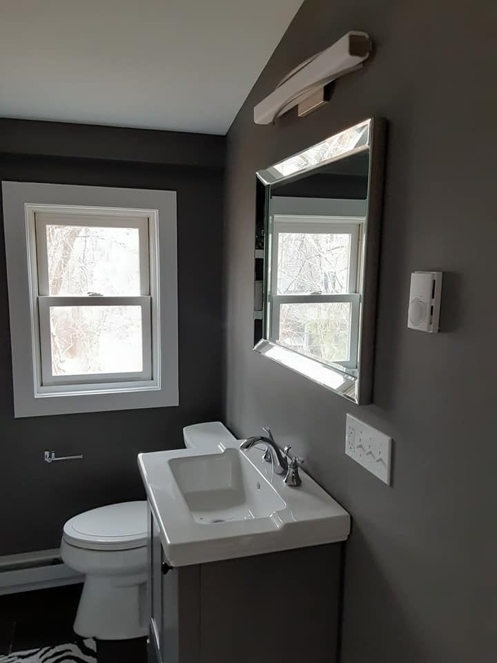 Small bathroom with gray walls, window, toilet, vanity sink, and mirror light fixture.