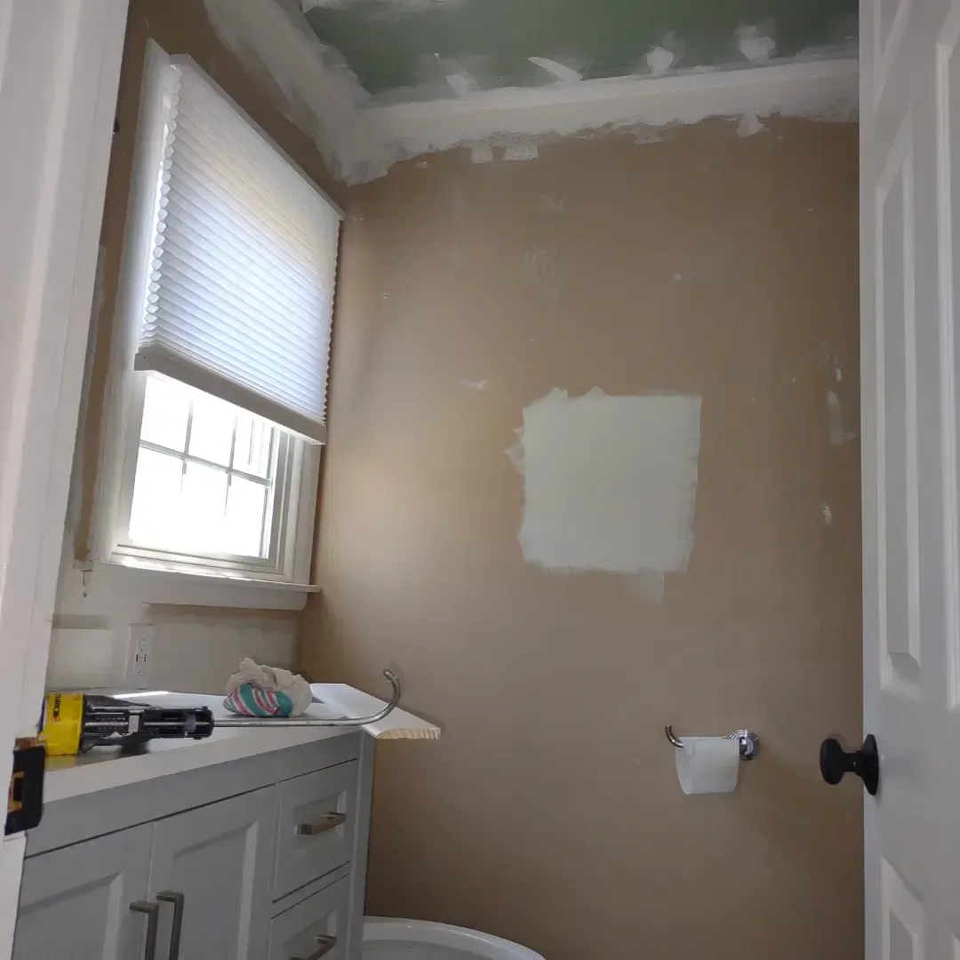Small bathroom mid-renovation with beige walls, patched drywall, window, sink cabinet, and toilet paper holder.