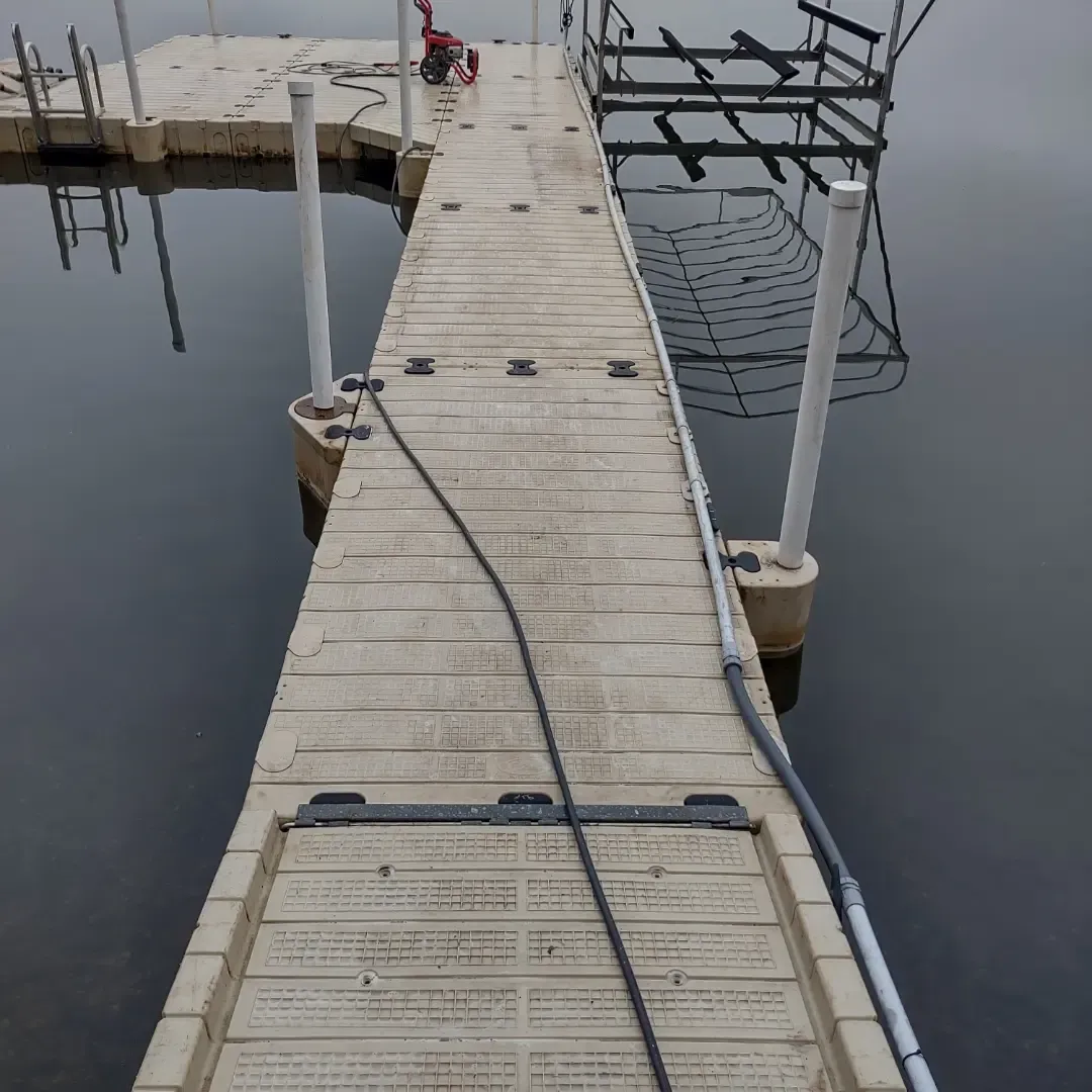 Wooden dock walkway over calm water with rope railings and a hose across the planks