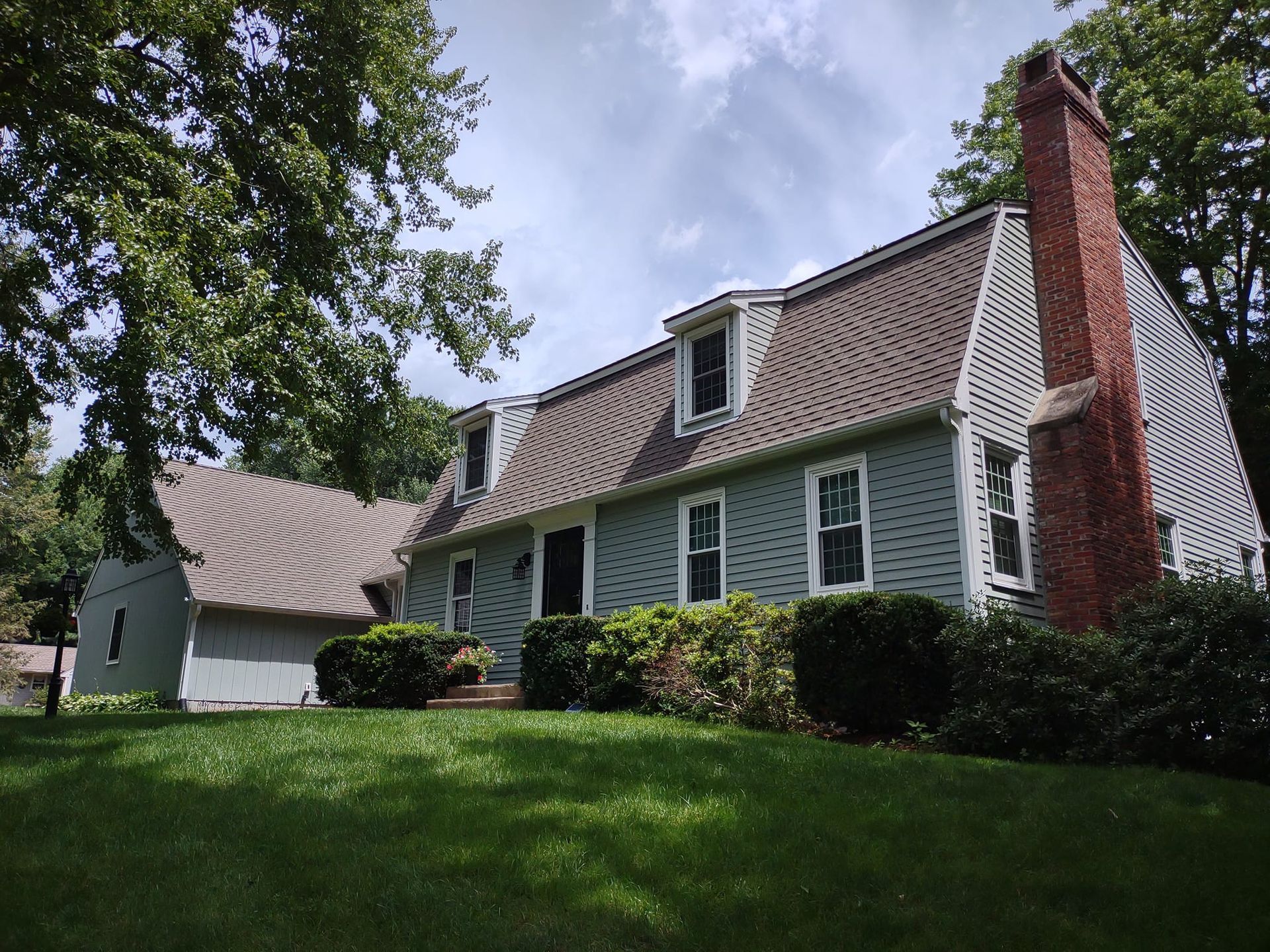 Green-shingled house with white trim, red brick chimney, and sloped roof amid a grassy yard and trees