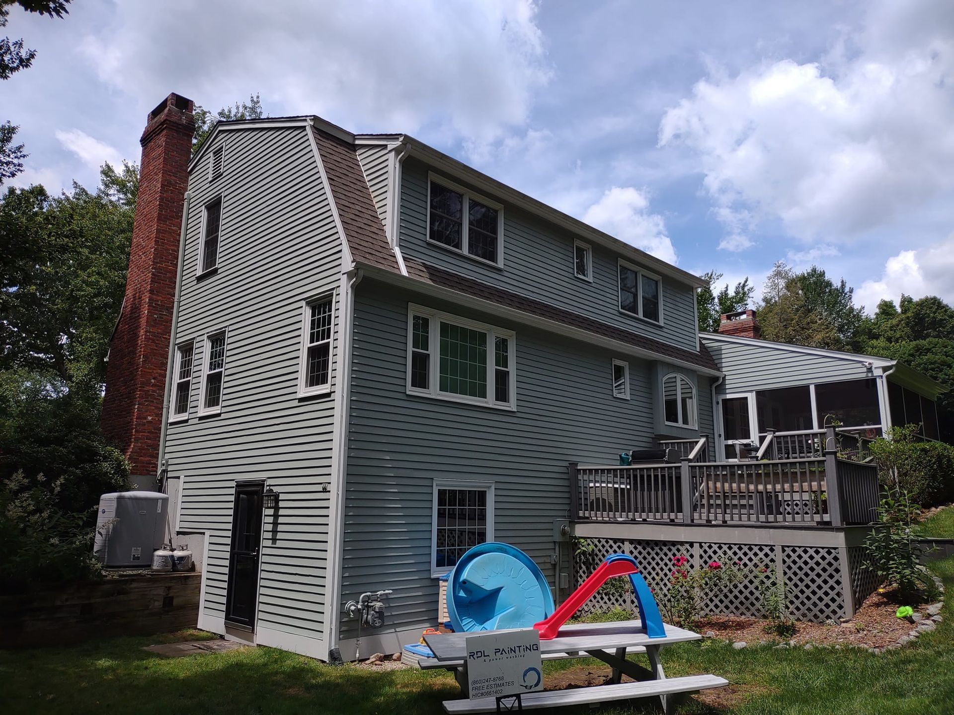 Gray house with white trim and deck in a backyard, with a brick chimney and a blue-and-red children's toy.