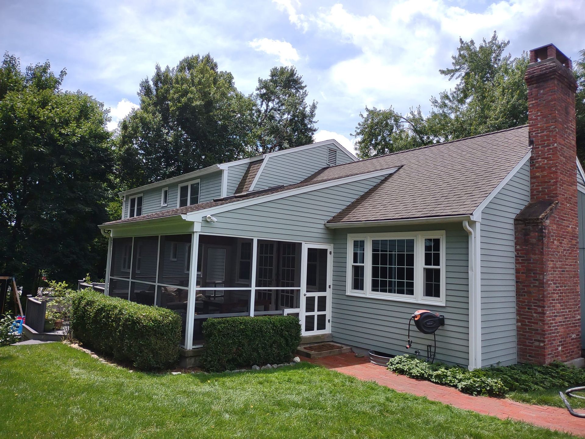 Suburban house with green siding, screened porch, brick chimney, and manicured lawn under a blue sky