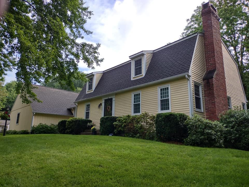 Suburban house with cream siding, dark roof, red brick chimney, and green lawn under trees