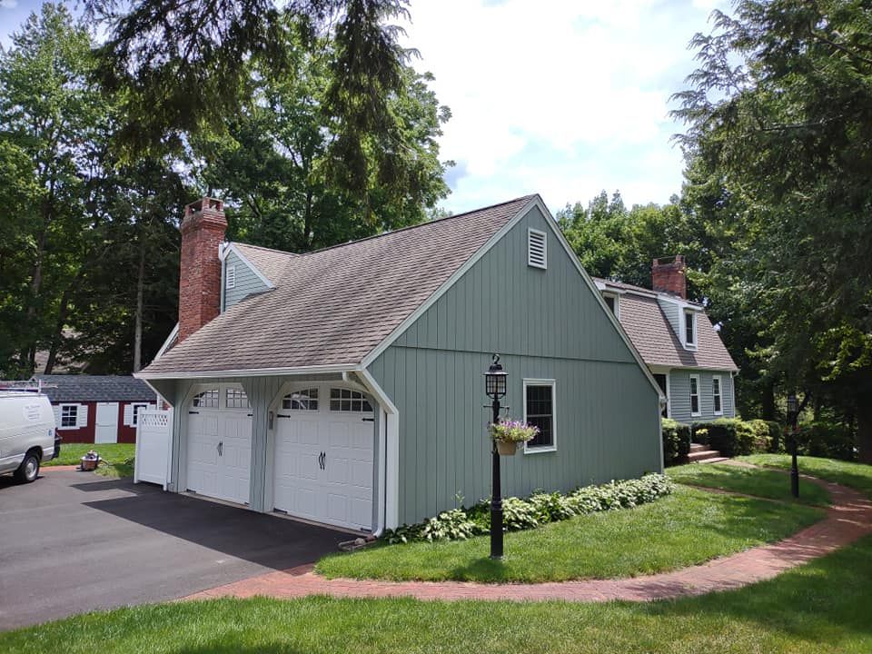 Green garage with white doors beside a house, bordered by a curved walkway and trees.