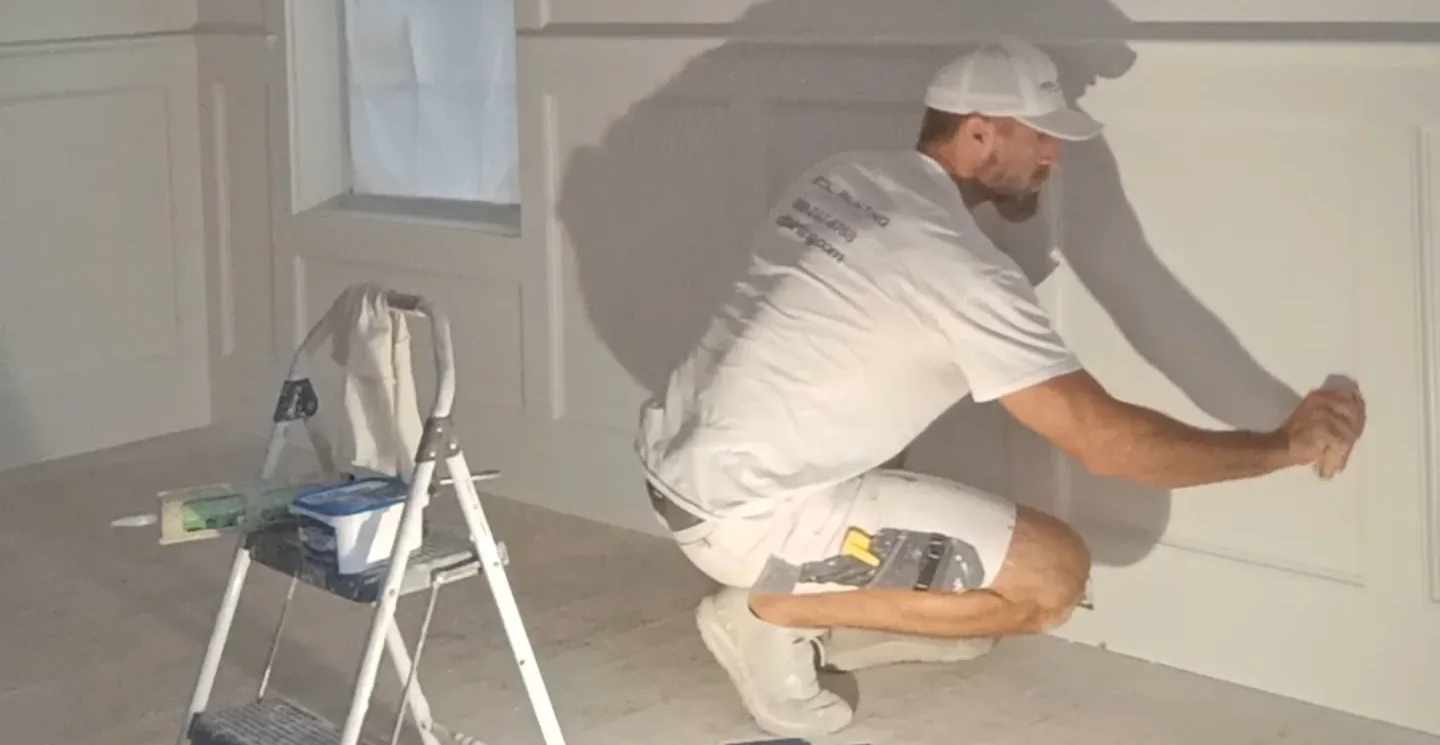 Painter kneeling on floor painting a white wall beside a step ladder and paint supplies