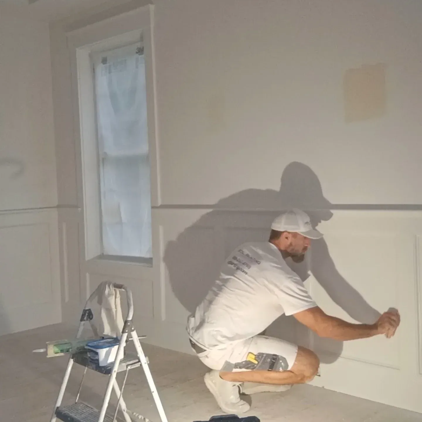 Painter crouching to paint white trim on a wall in an empty room, with a step ladder nearby.