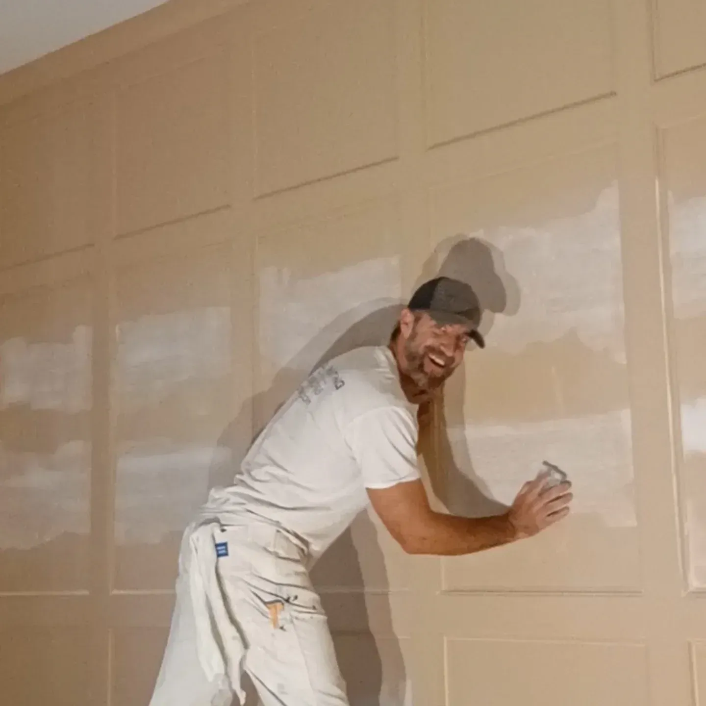 Painter in white clothes and cap applying paint to a beige paneled wall.