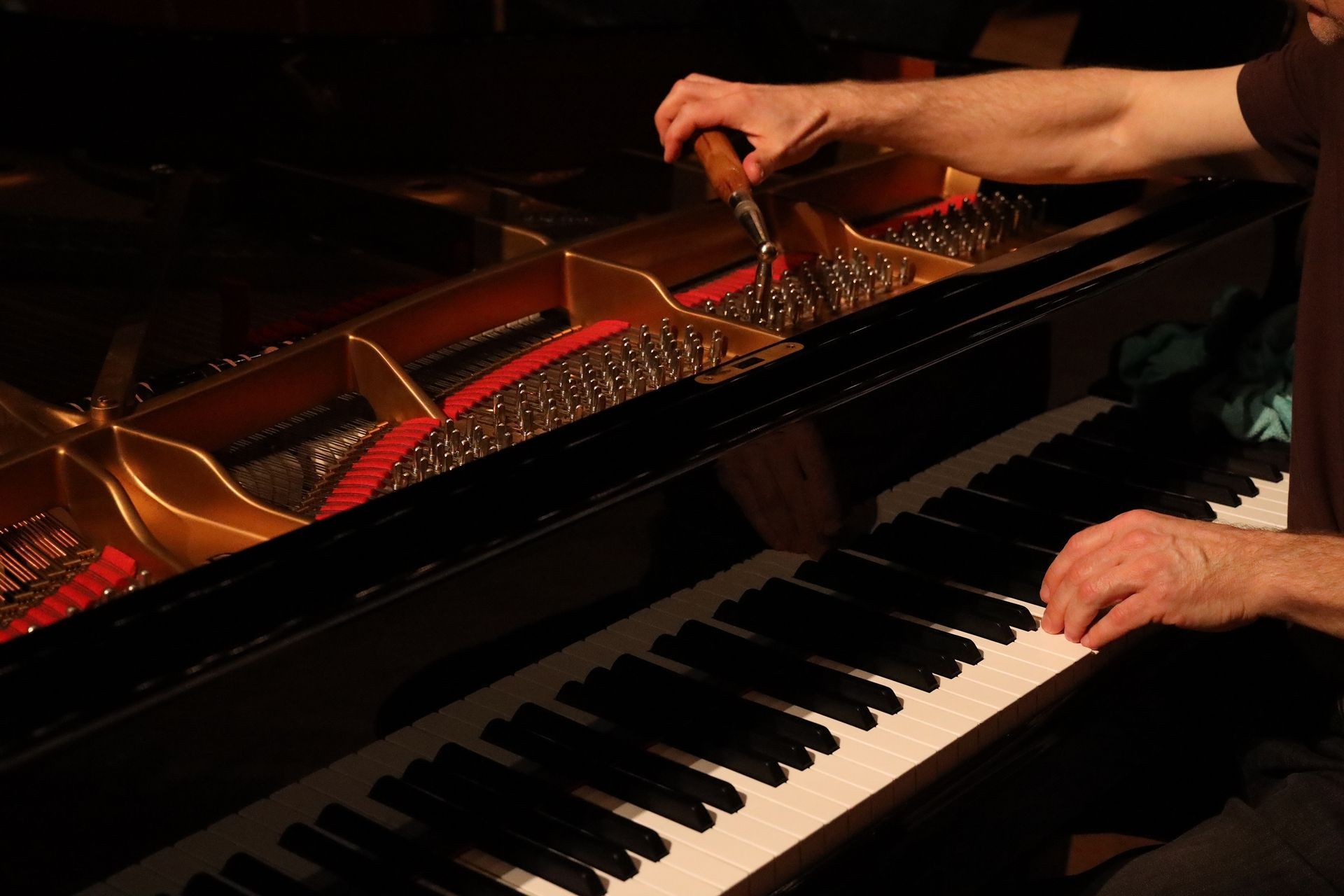 Hands playing a grand piano with the lid open, showing strings and a warm-lit interior.