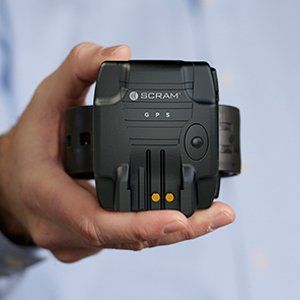 Hand holding a black SCRAM GPS ankle monitor against a light blue shirt