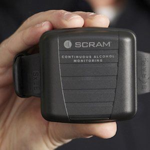 Hand holding a black SCRAM continuous alcohol monitoring ankle monitor bracelet