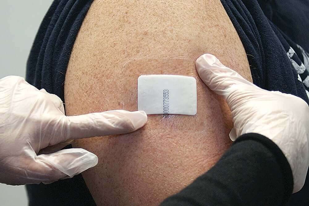 Gloved hands apply a small adhesive patch to a person's upper arm