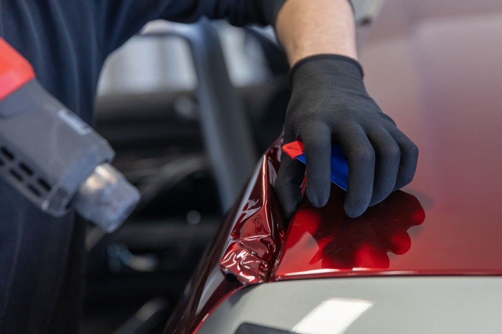 Gloved hand applying red vinyl wrap to a car panel with a heat gun nearby