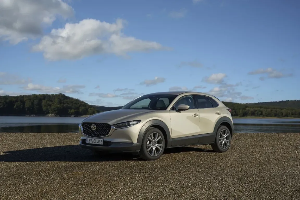 Ein hellbeiger Mazda CX-30 SUV parkt auf einem Kieselstrand neben einem See unter einem teilweise bewölkten blauen Himmel.