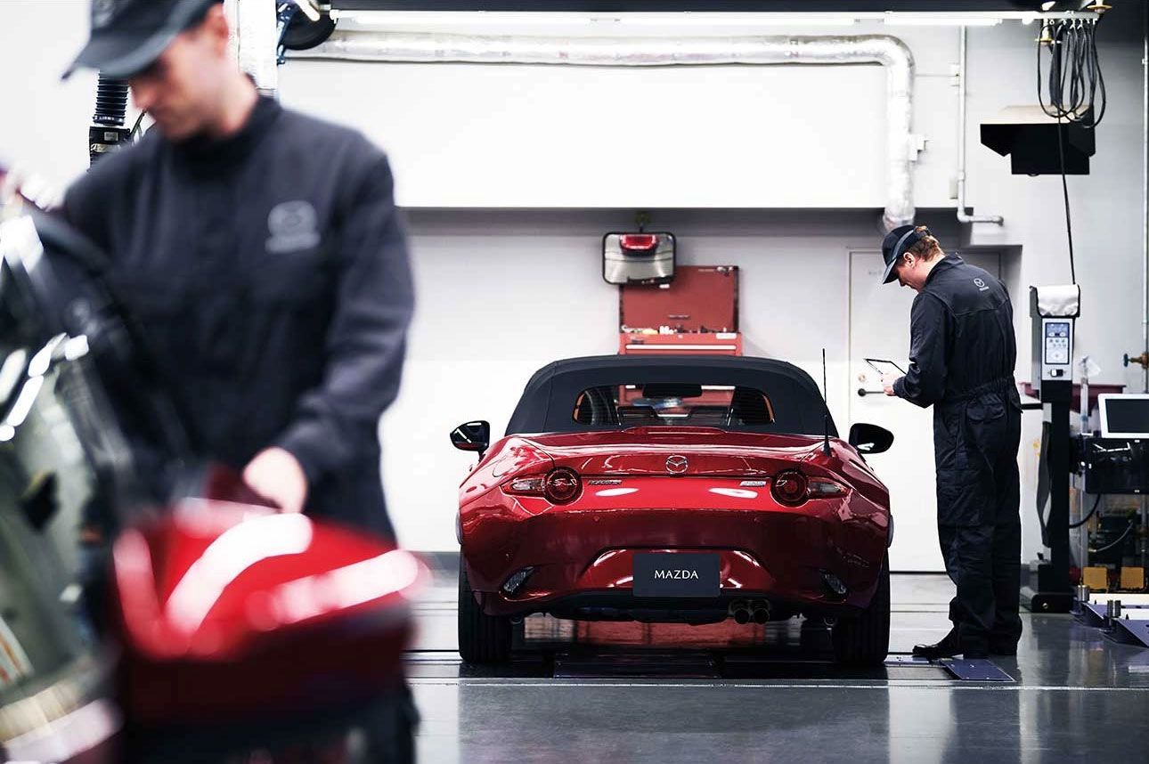 Zwei Techniker in dunklen Overalls arbeiten in einer hellen Werkstatt an einem glänzenden roten Cabrio-Sportwagen.