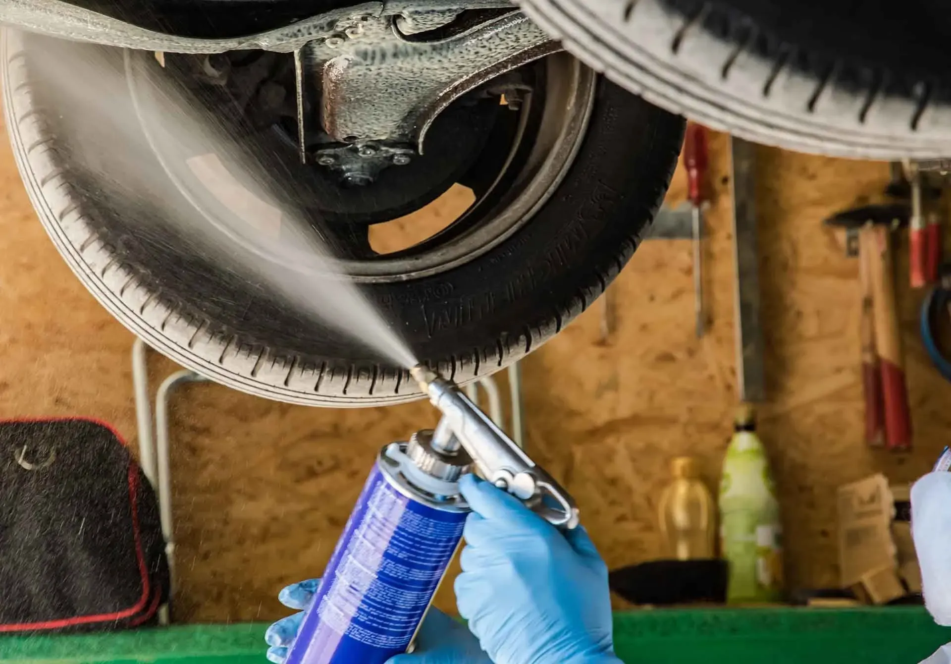 Eine Person mit blauen Handschuhen besprüht in einer Garage die Unterseite eines Autoreifens mit einer Sprühdose.