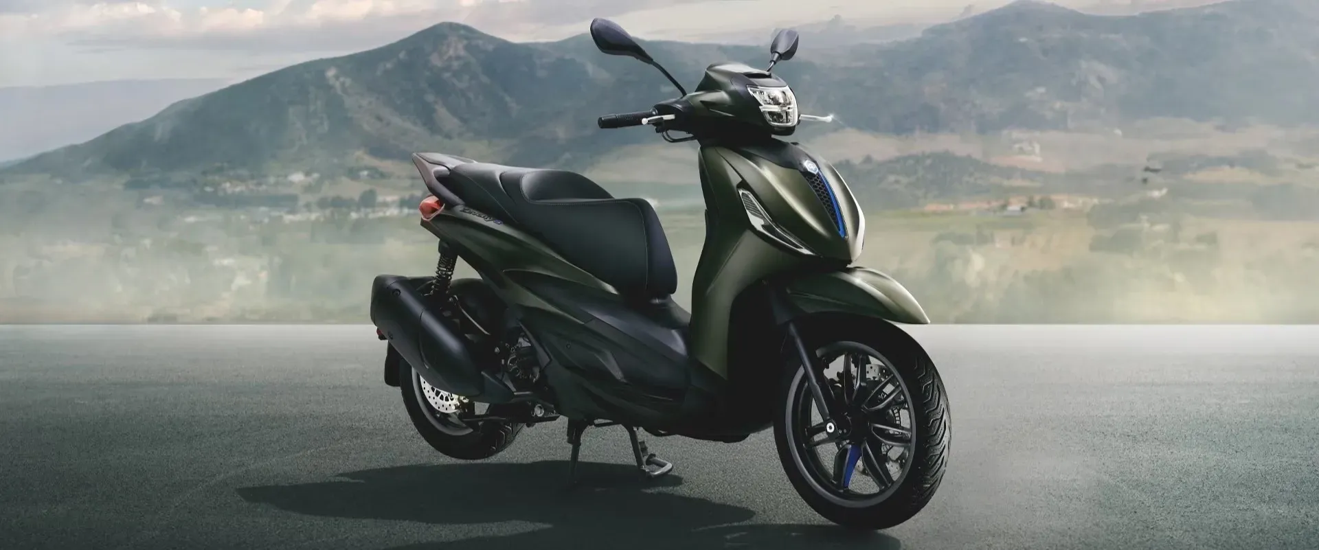 Ein olivgrüner Piaggio Beverly-Roller parkt am Straßenrand vor einer Bergkulisse.
