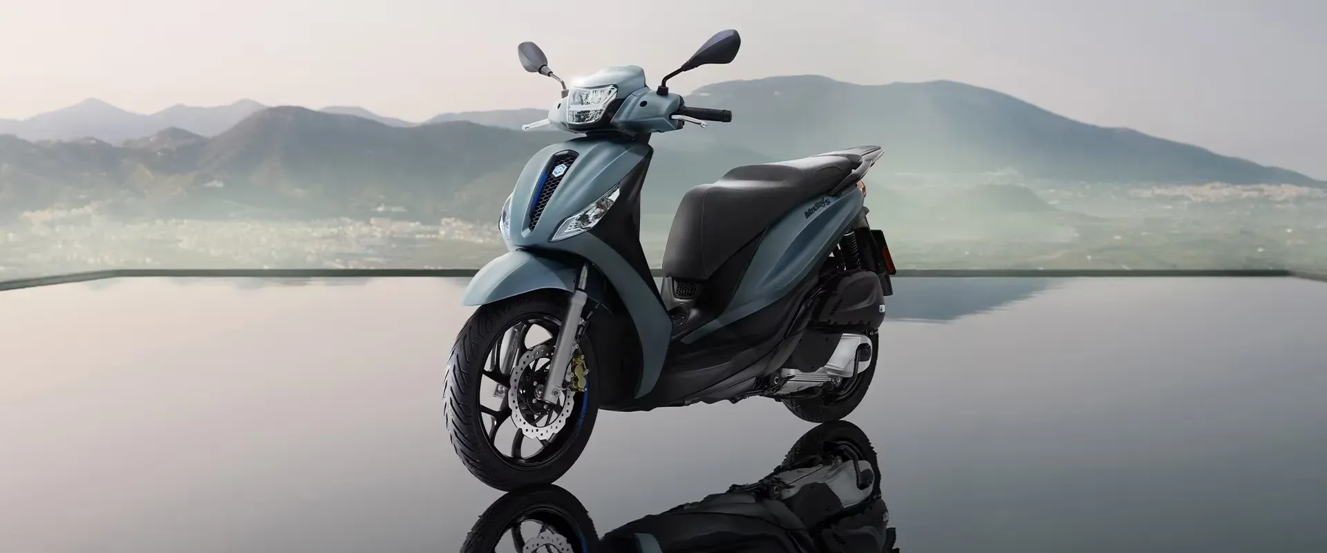 Ein mattblauer Piaggio Medley-Roller parkt auf einer spiegelnden Fläche vor einer Berglandschaft.