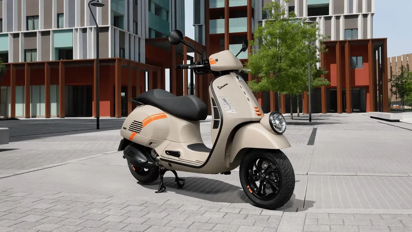 Ein beigefarbener Vespa-Roller mit orangefarbenen Rennstreifen parkt auf einem gepflasterten Platz vor modernen Gebäuden.