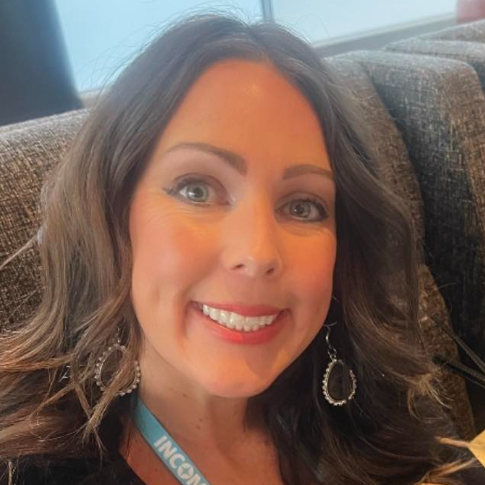 Smiling woman with dark hair and earrings on a couch, wearing a blue lanyard, in a bright room