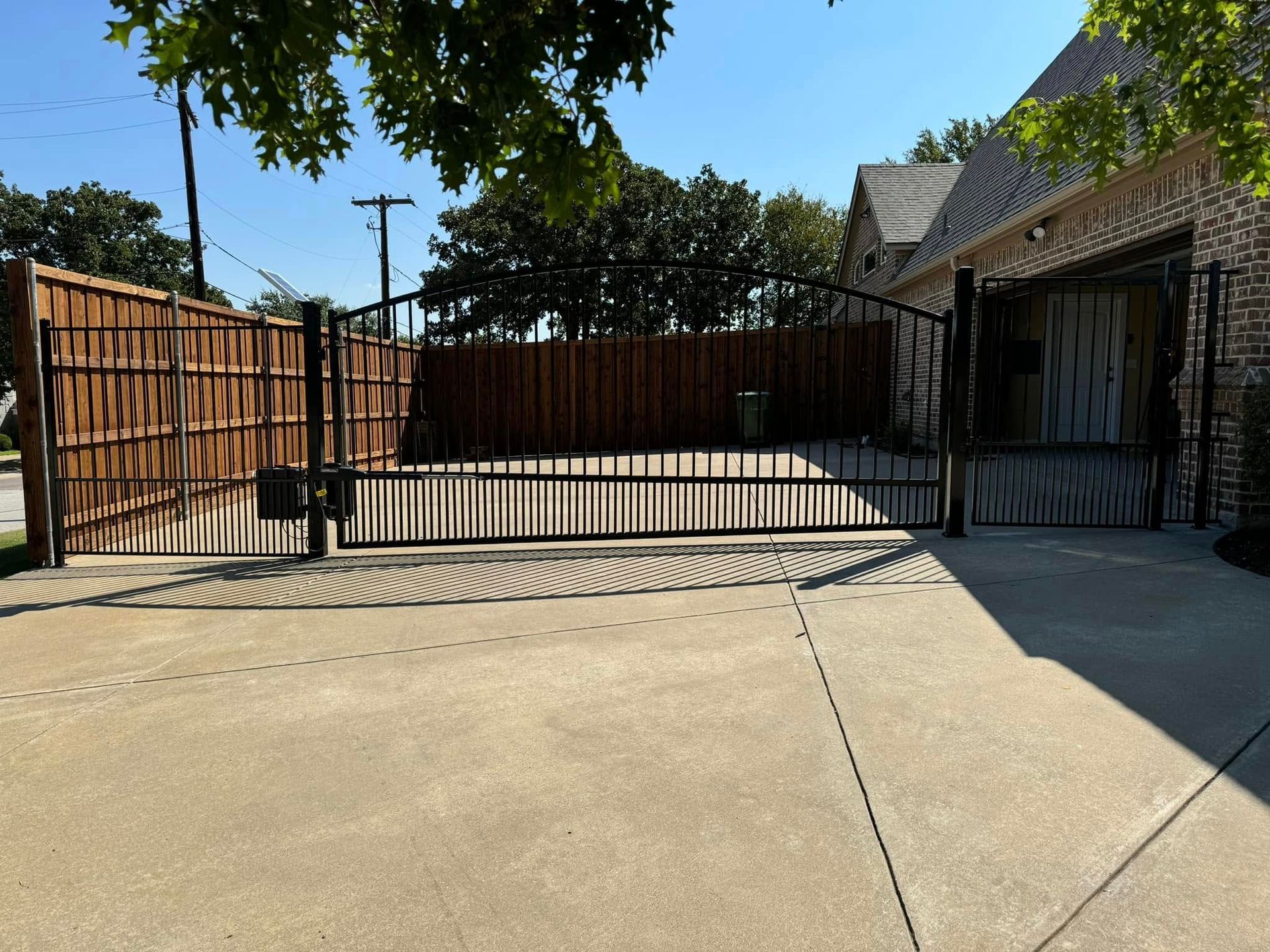 A black metal gate in front of a driveway, flanked by wooden and brick structures, on a sunny day.