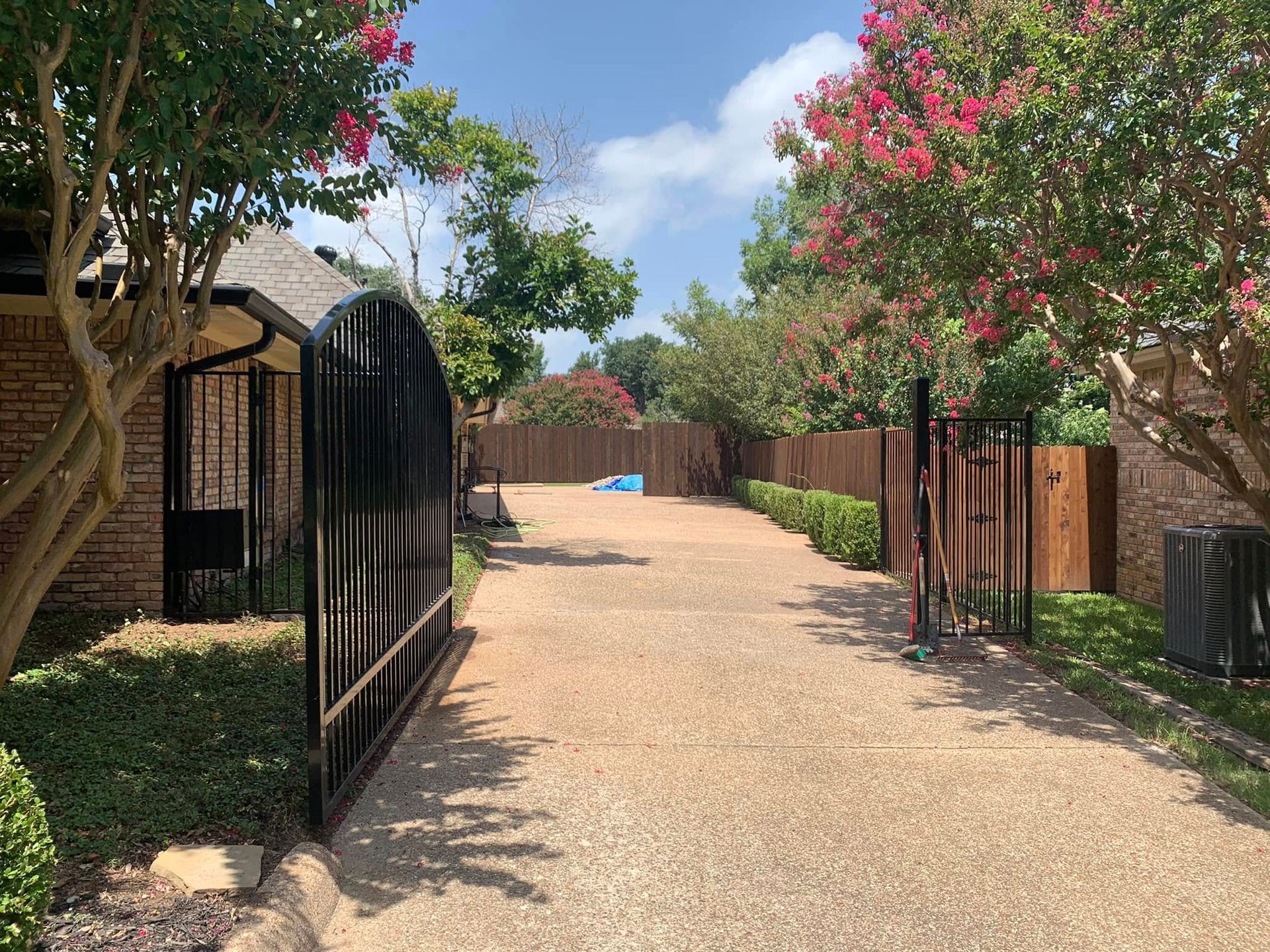 Driveway leading to a house, flanked by trees and an open black metal gate; gravel surface, bright sunlight.