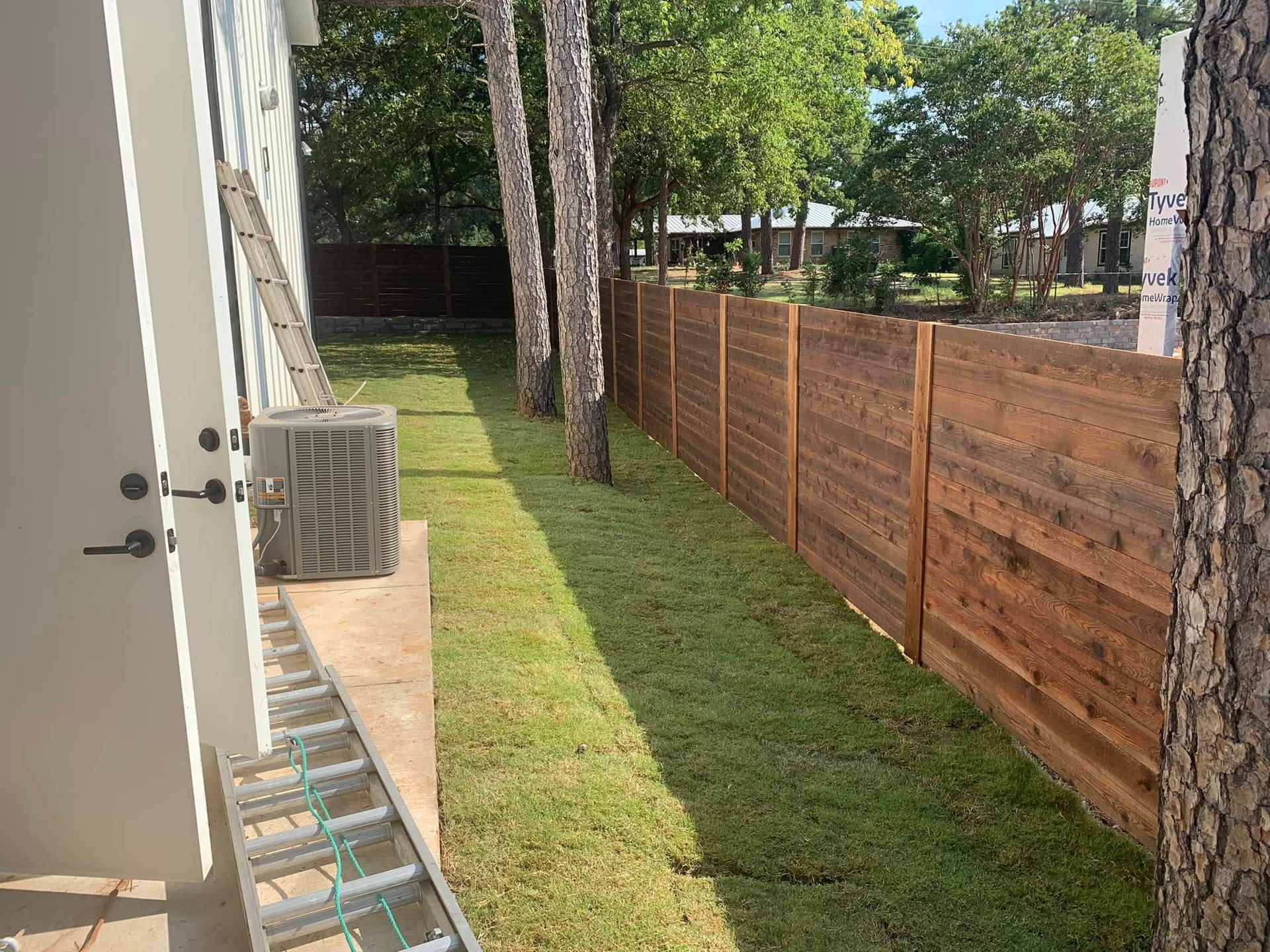 Backyard with wooden fence, grass, air conditioner, and trees.