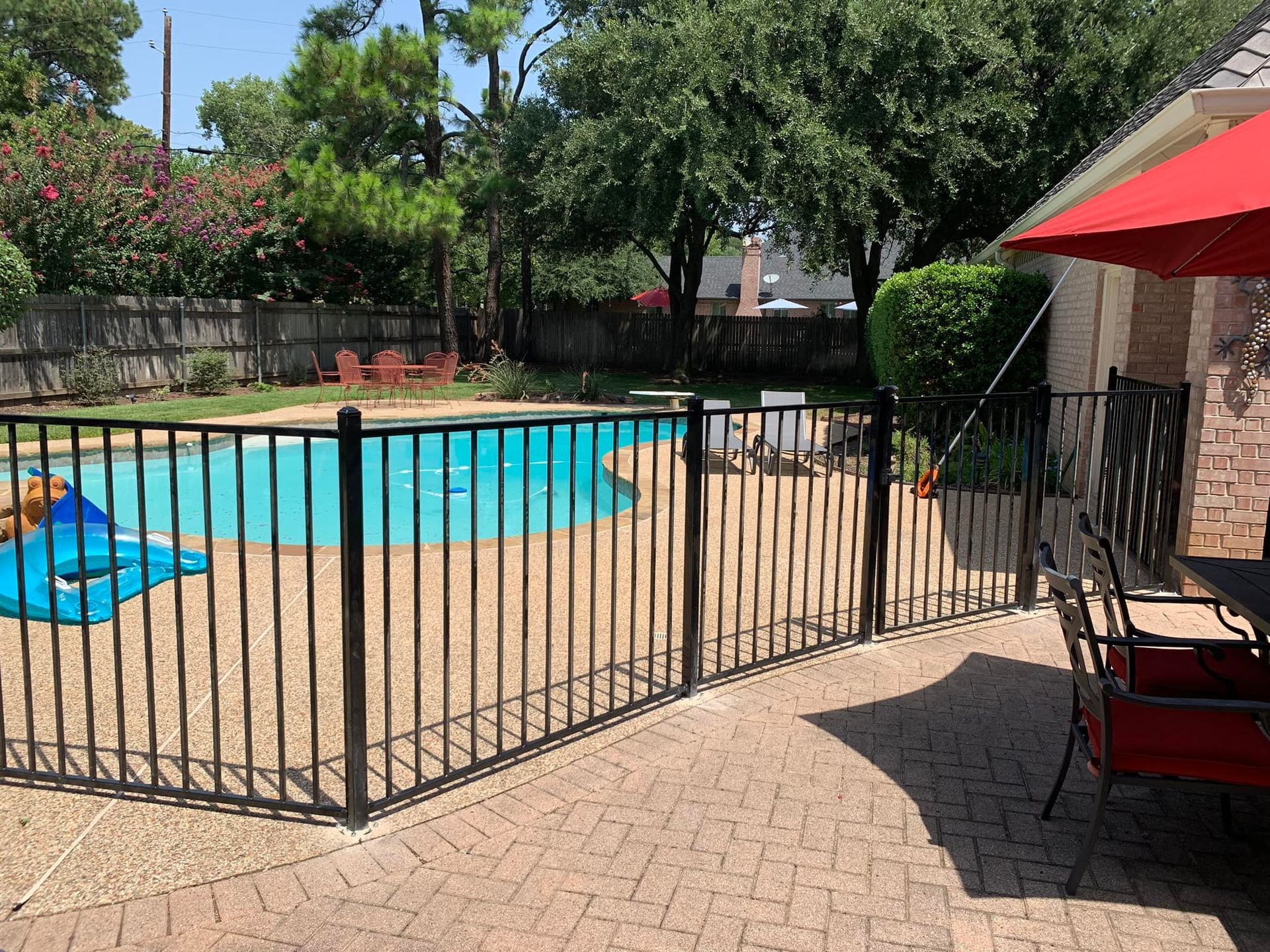 Black fenced pool, patio with table and chairs, red umbrella, and lush backyard greenery.