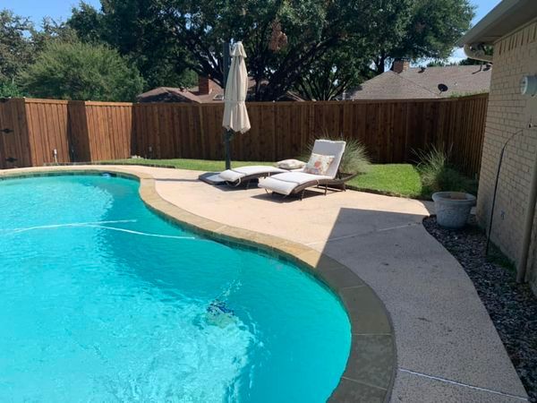 Poolside scene with a pool, lounge chairs, and a closed umbrella next to a fence and lawn.