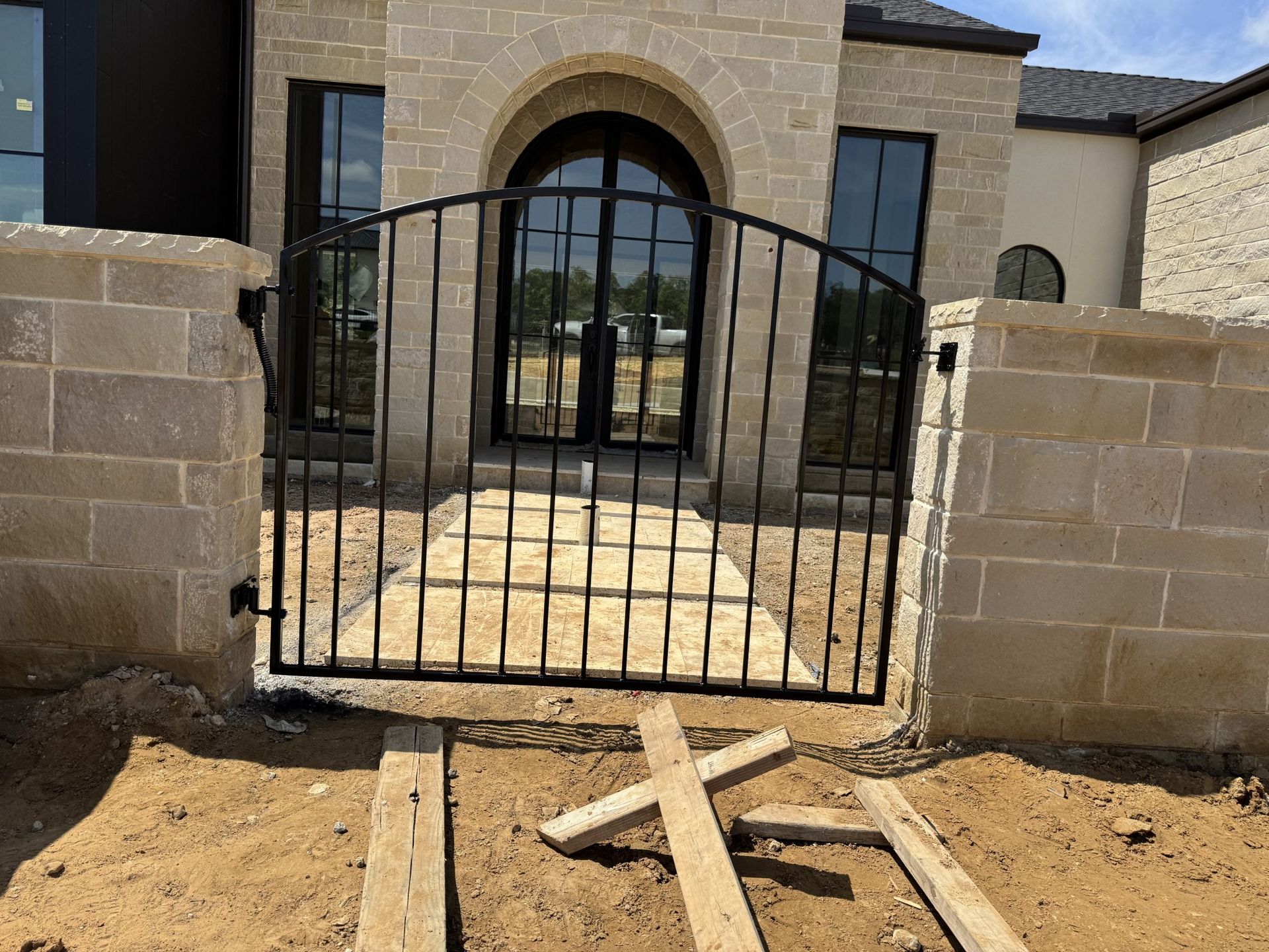 Black iron gate in front of a beige stone house with a arched doorway.