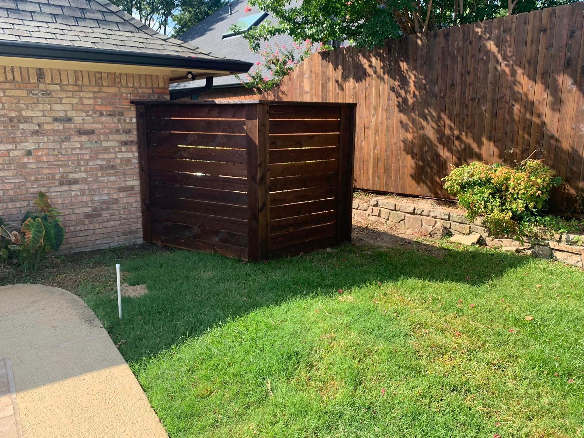 Wooden trash enclosure in a backyard, beside a brick wall and a wooden fence.