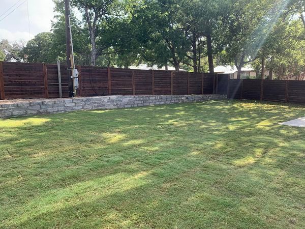 Green backyard with brown fence, retaining wall, and trees under a sunny sky.