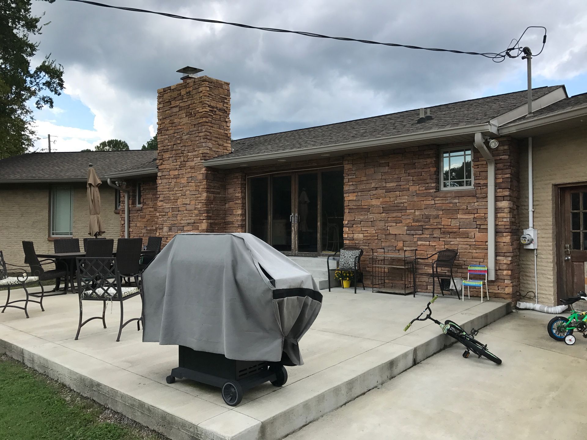Backyard patio with covered grill, brick fireplace, outdoor chairs, and toys beside a house