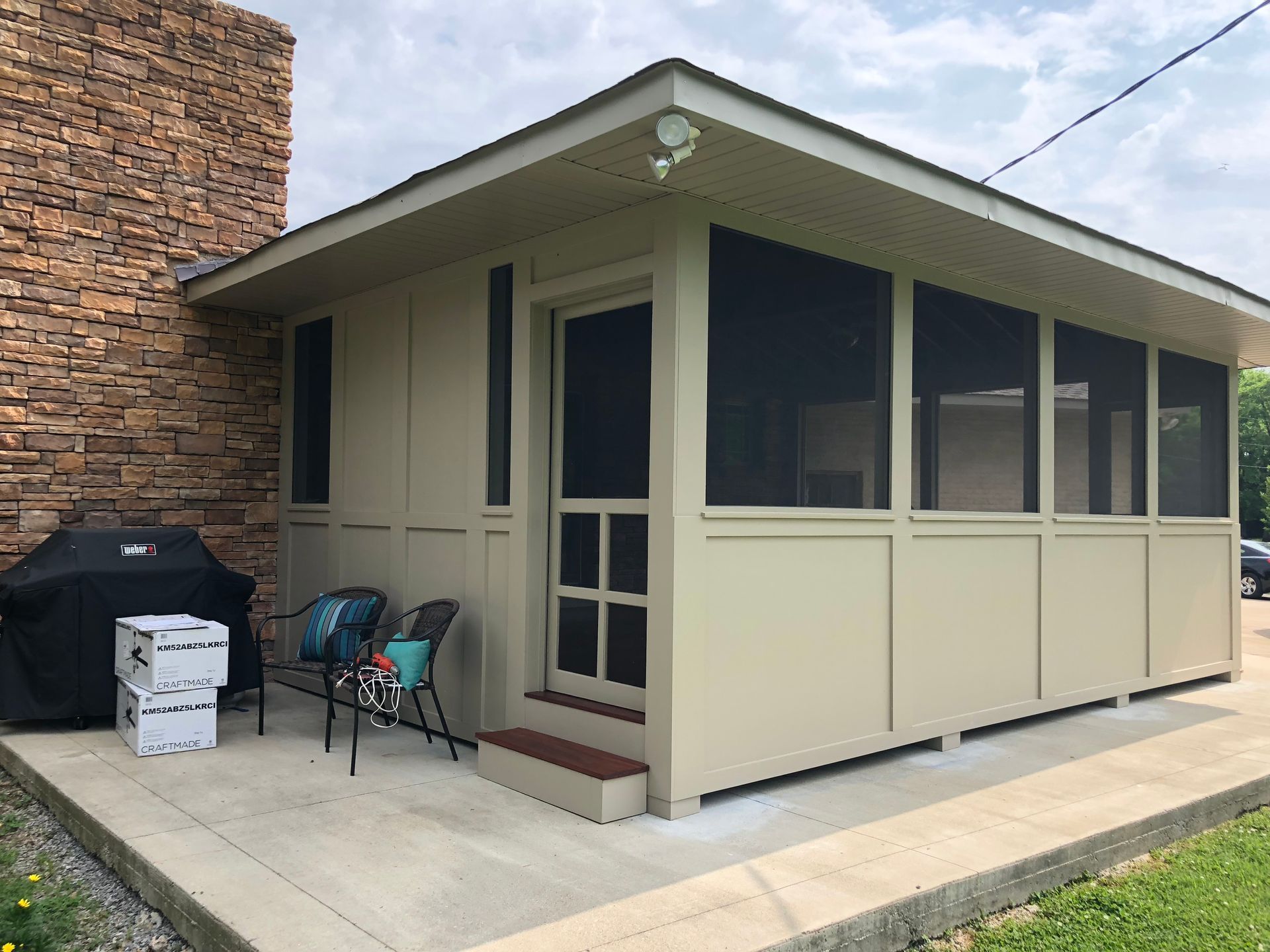 Beige screened porch with a glass door beside a brick house and patio chairs on a concrete slab.