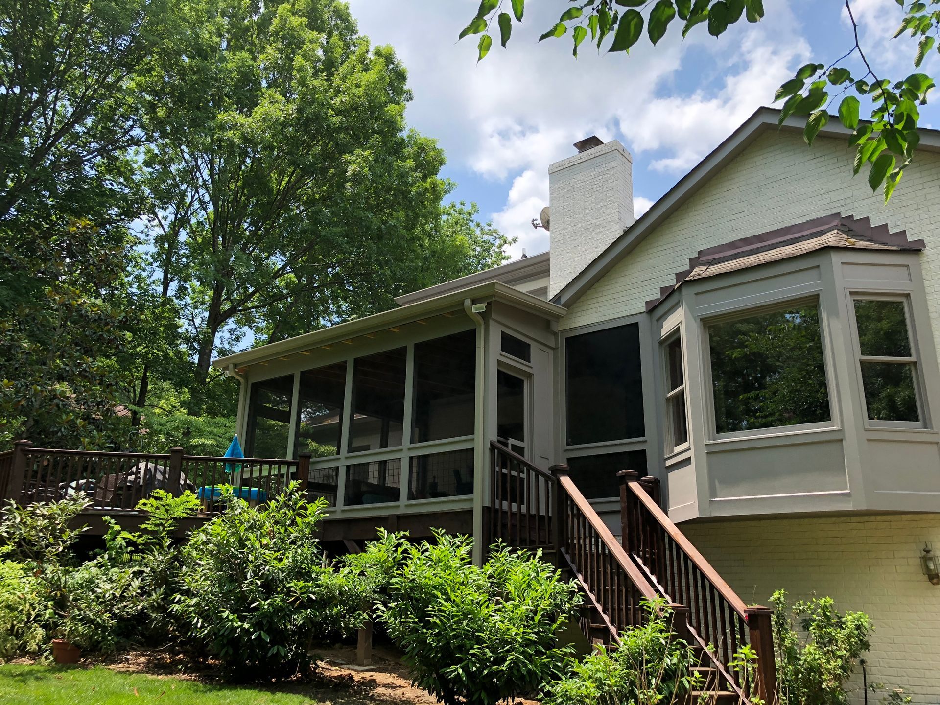 House with screened porch and bay window surrounded by lush green trees and shrubs