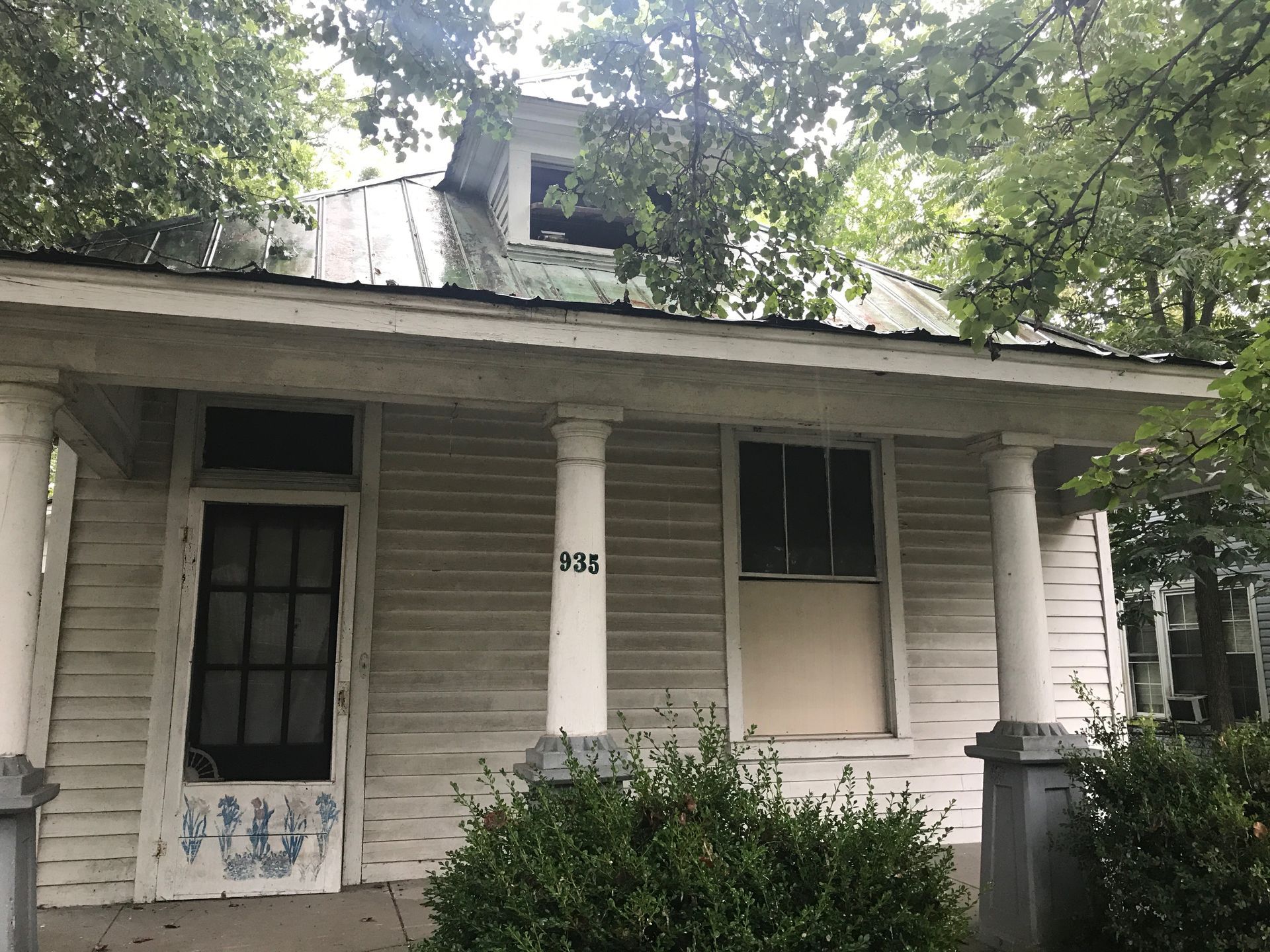 Small weathered house with white porch columns and overgrown bushes under trees.