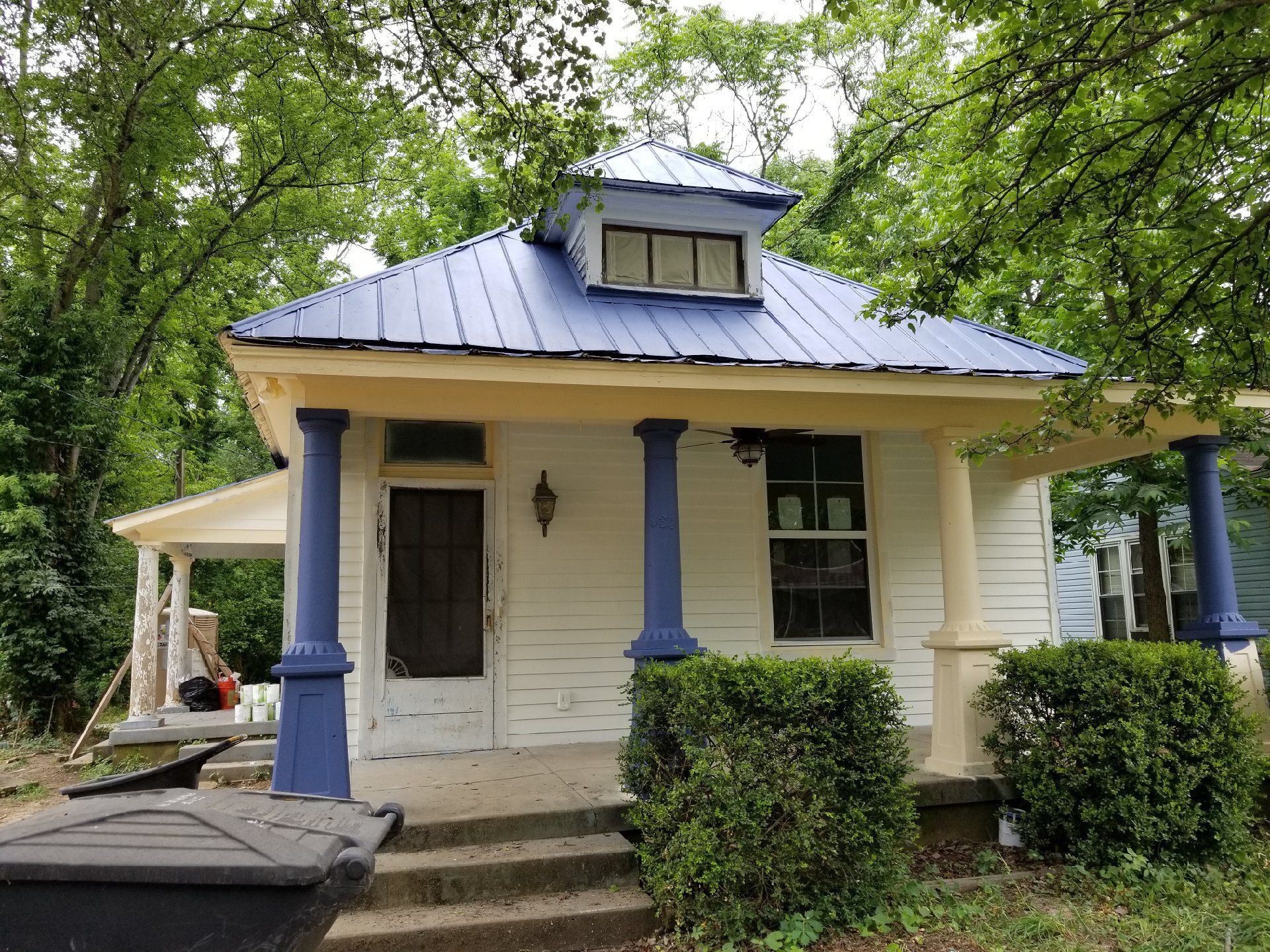 Small cream cottage with a blue metal roof, front steps, and porch surrounded by trees and bushes