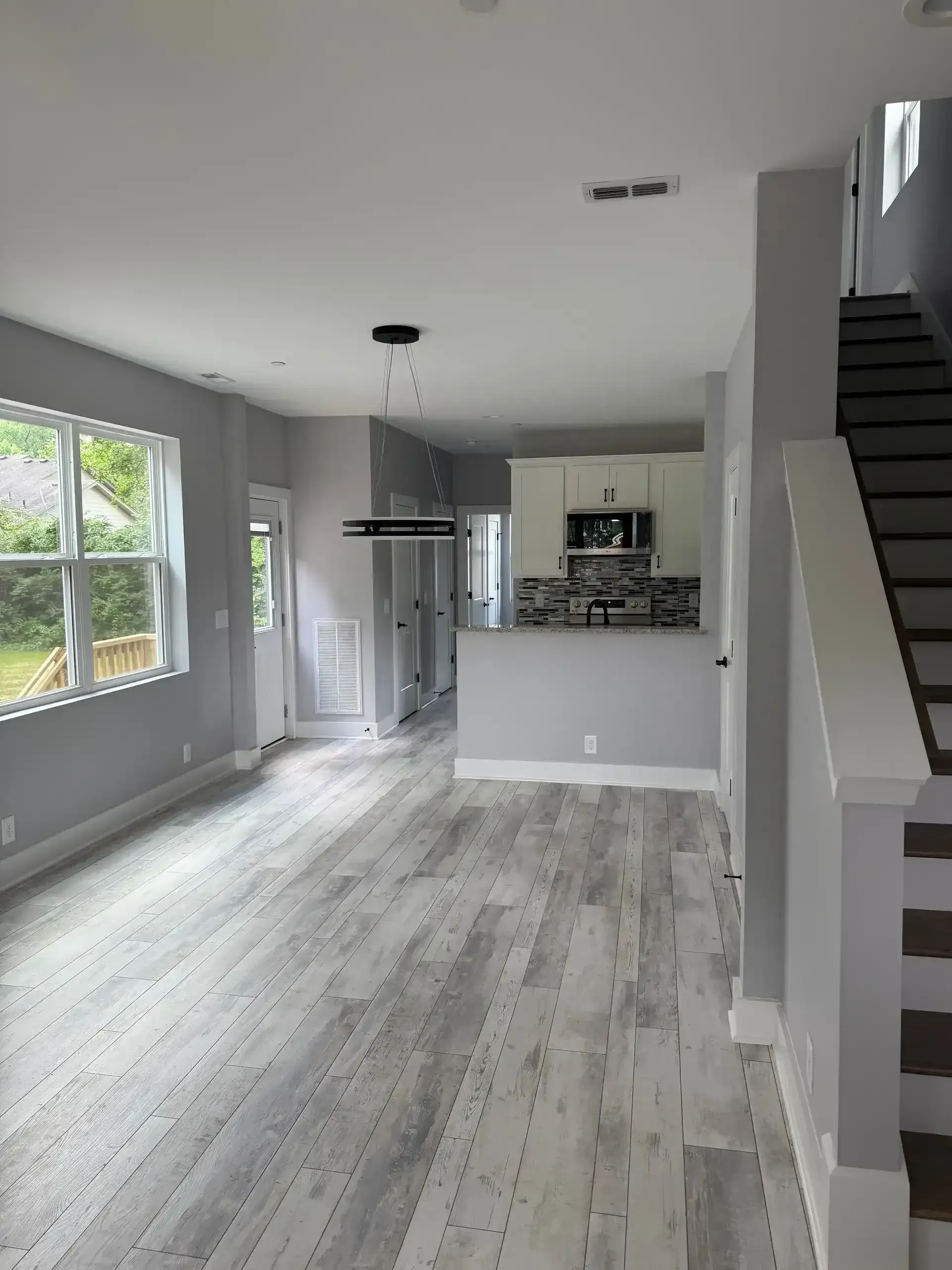 Empty gray modern living room with light wood floors, kitchen pass-through, and staircase to the right