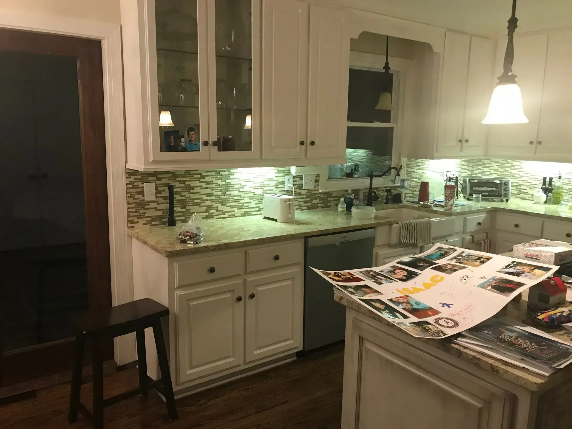 Dim kitchen with white cabinets, green backsplash, and papers spread on the island counter.