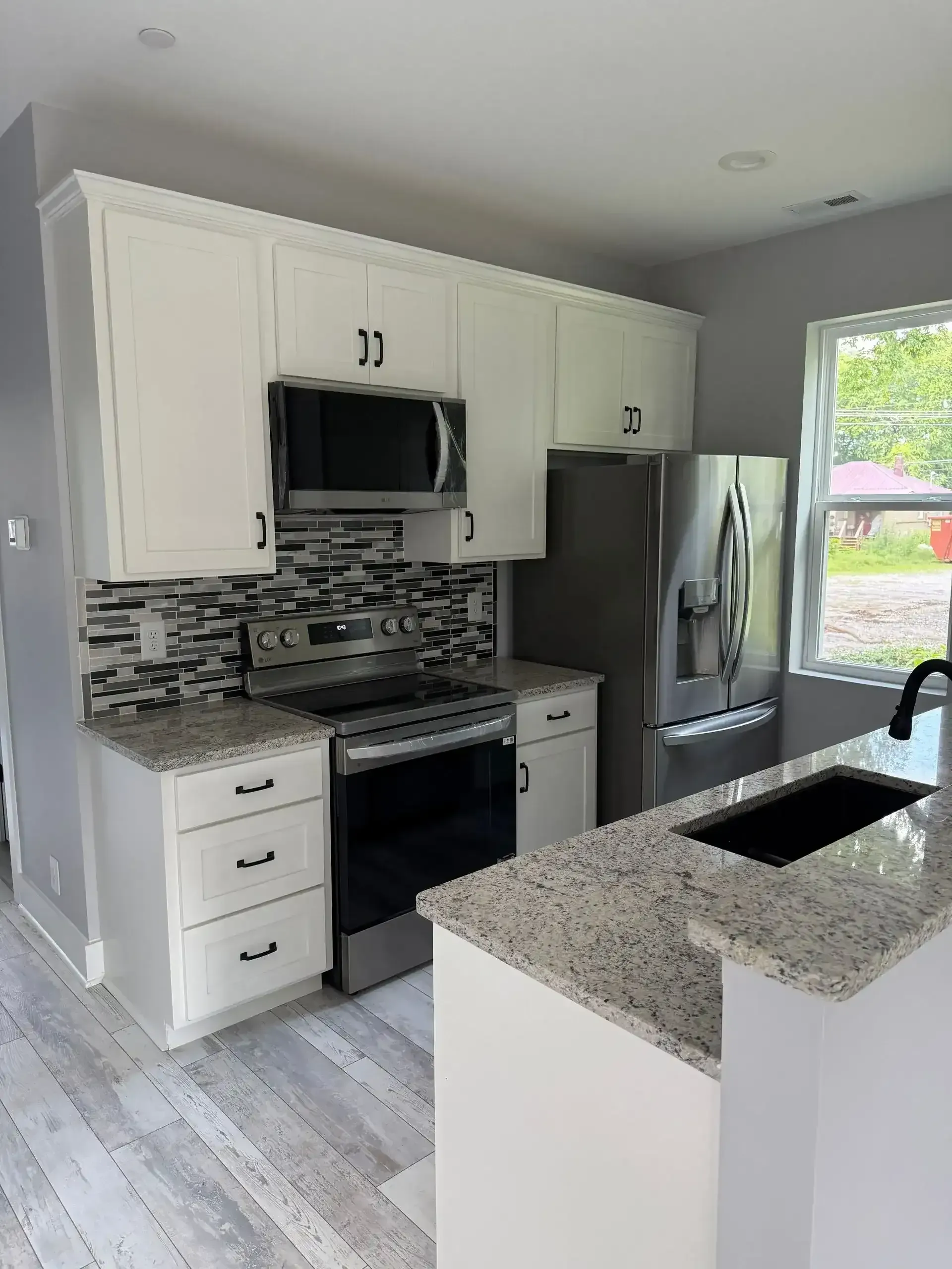 Modern white kitchen with stainless steel appliances, gray mosaic backsplash, granite island, and a window by the sink