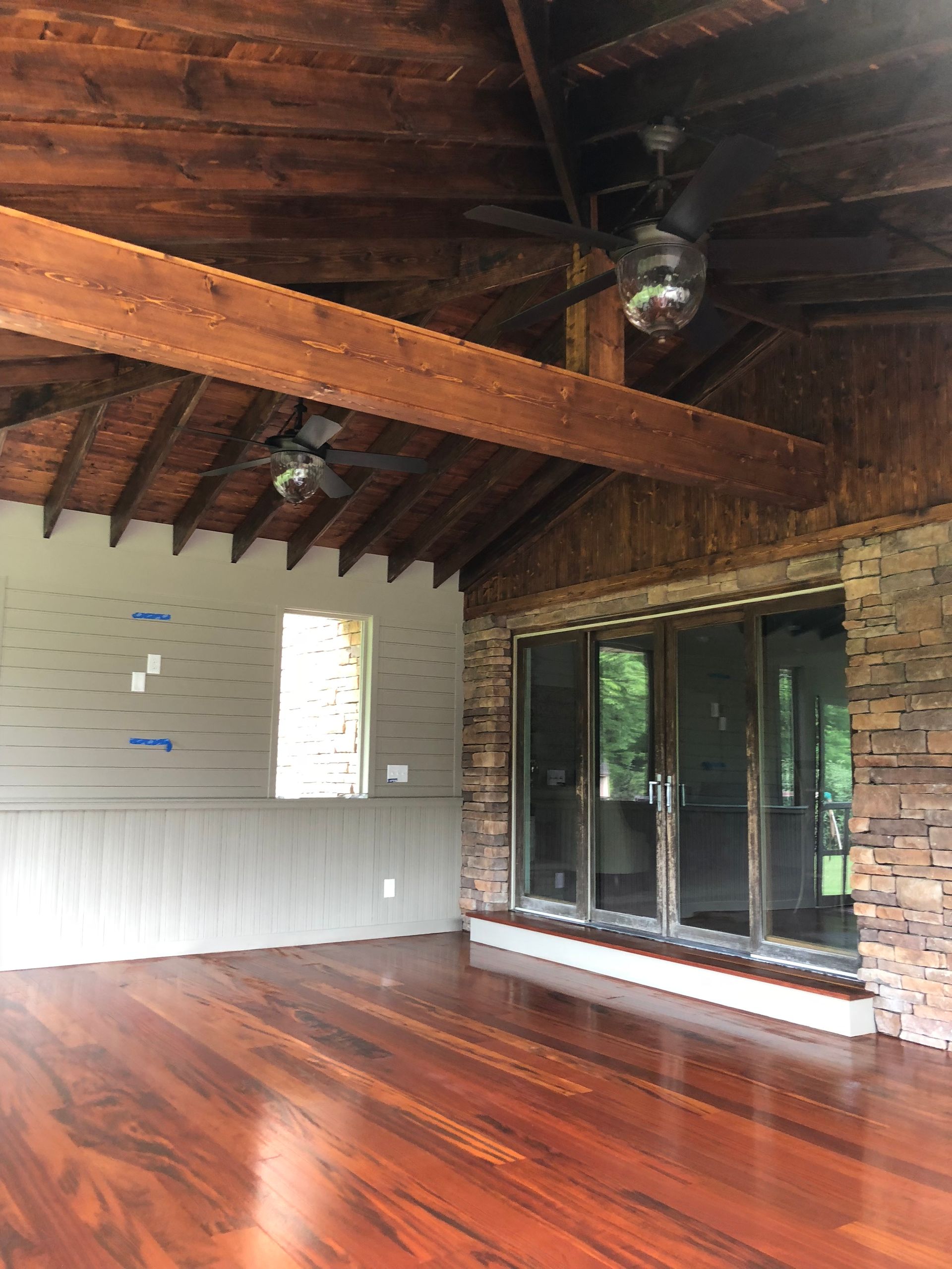 Empty rustic room with polished wooden floor, exposed beams, and large sliding glass doors opening to greenery