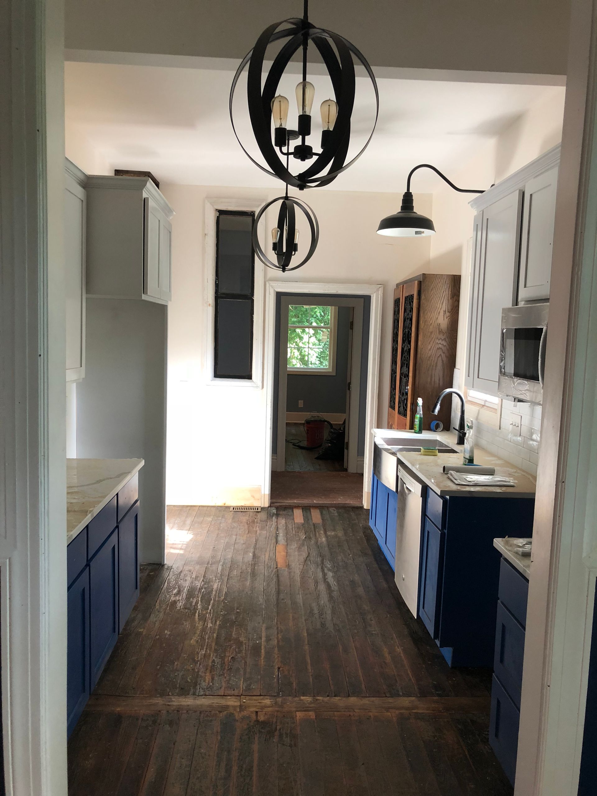 Narrow kitchen hallway with blue cabinets, wood floor, and hanging black pendant lights leading to a back room