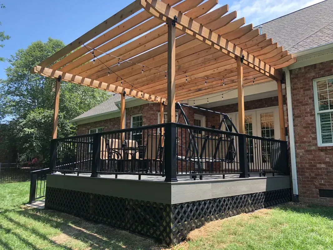 Covered backyard deck with black railings and a wooden pergola beside a brick house