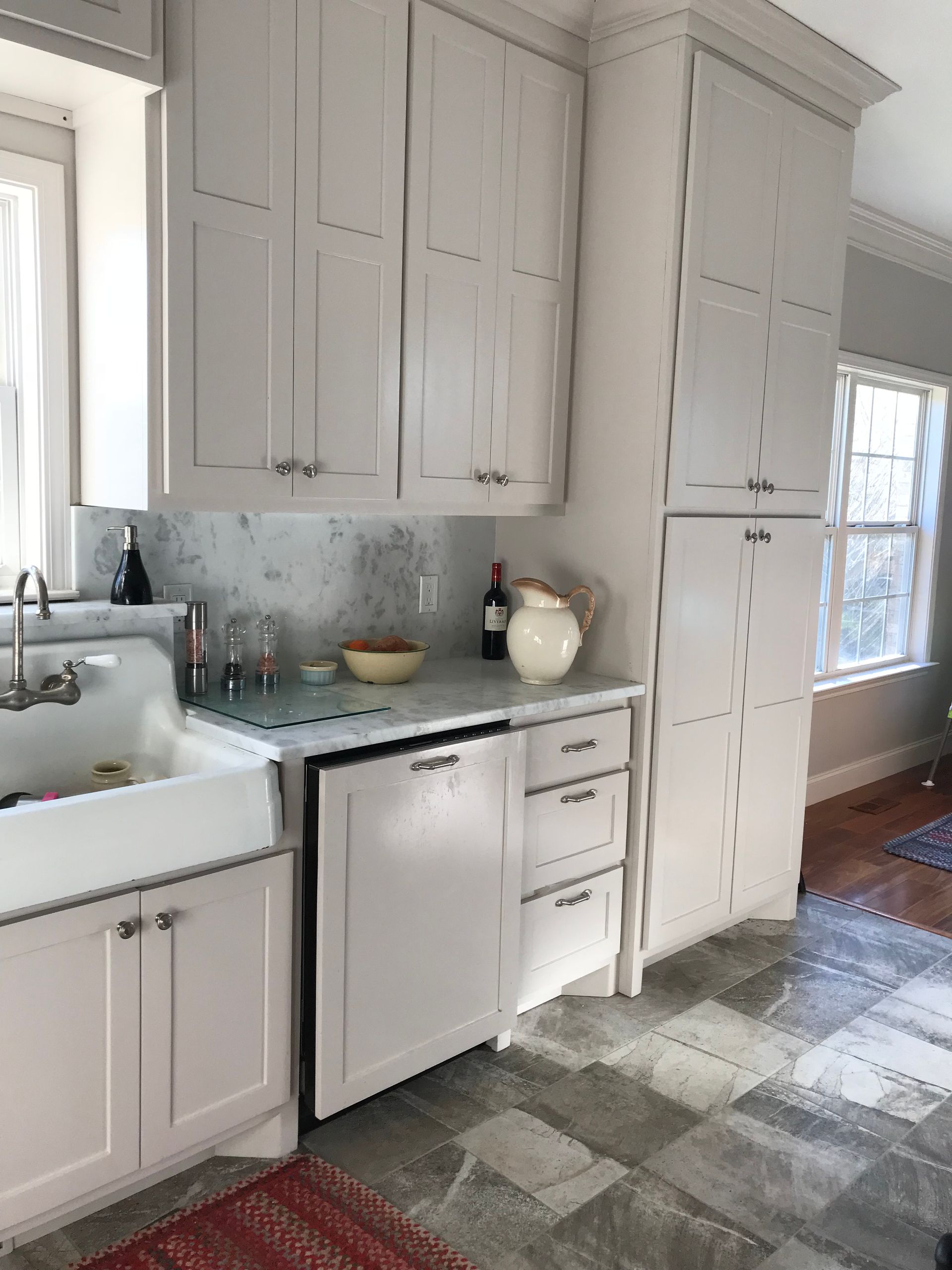 Bright kitchen with white cabinets, sink, dishwasher, and a checkered rug by a sunlit window.