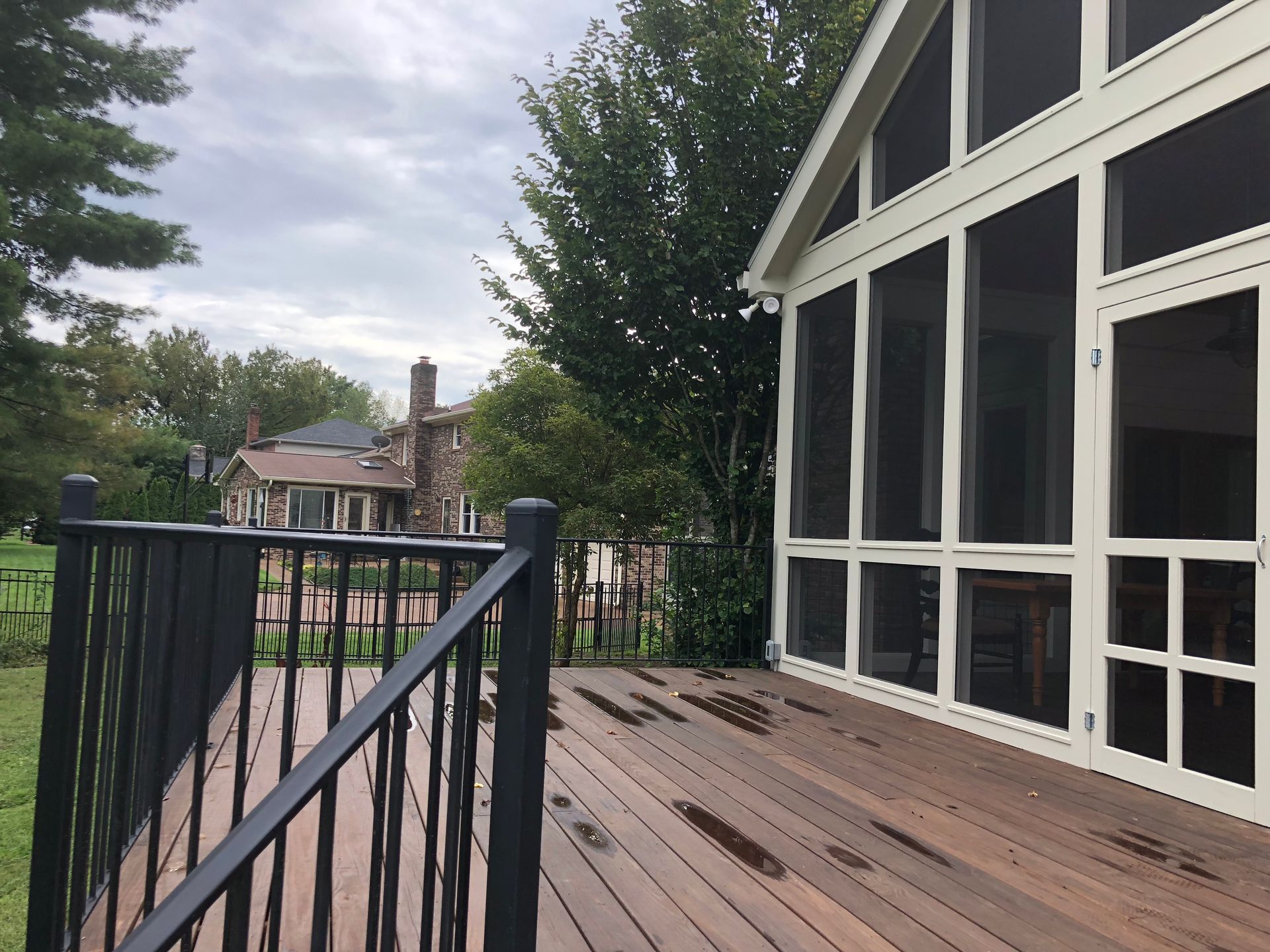 Wooden deck with black railing beside a white house, overlooking a leafy backyard and neighboring homes