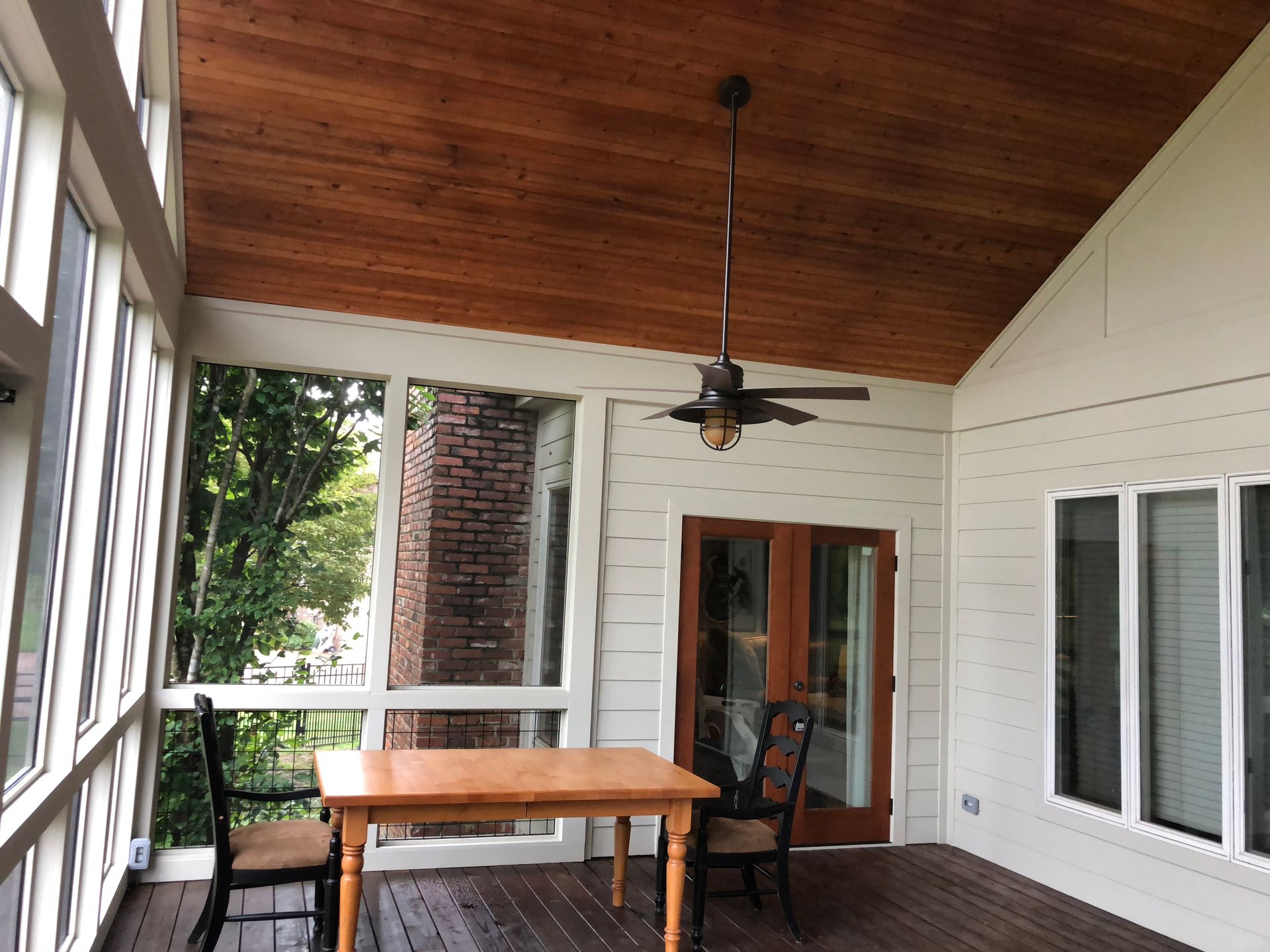 Covered patio with wood ceiling, ceiling fan, table, and sliding glass door beside white exterior windows