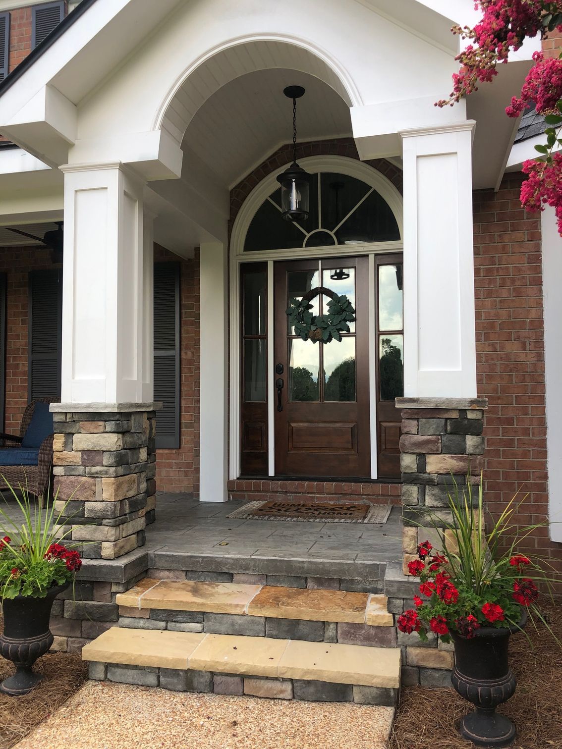 Front porch with brick steps, white arches, wooden double doors, and potted red flowers.